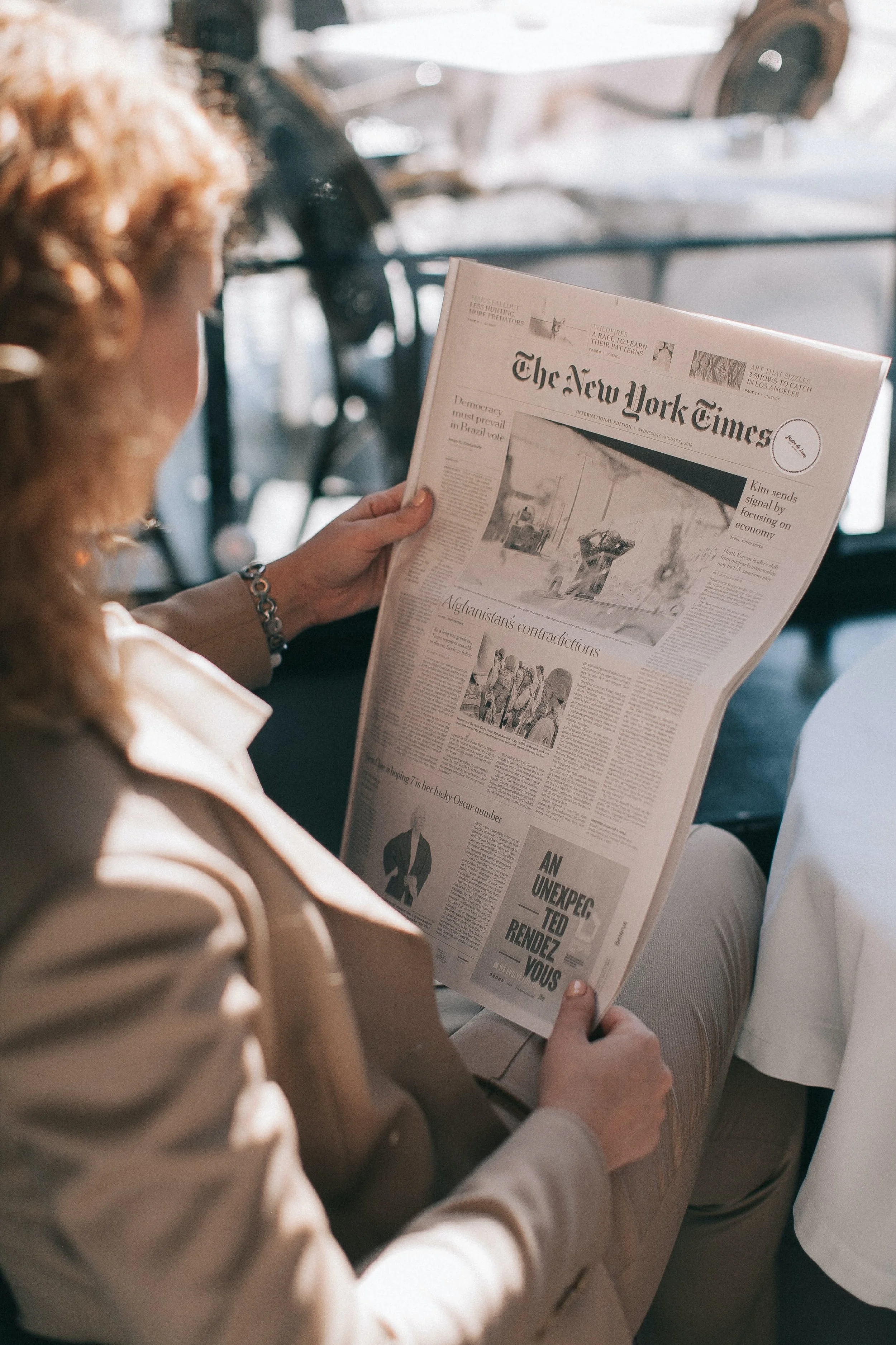 Person reading a newspaper, seated in a vehicle, with a blurred background showing part of a car and window.
