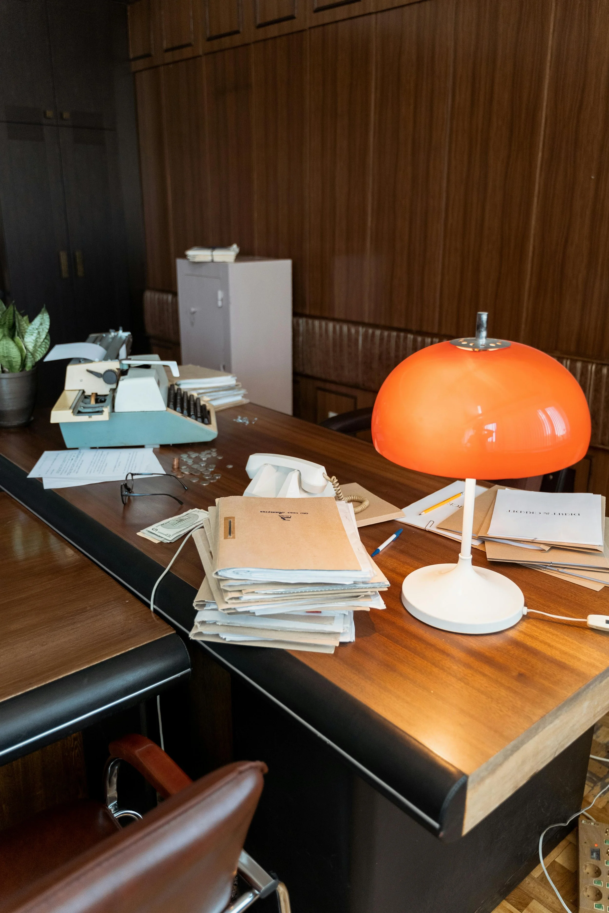 An office desk cluttered with papers, a vintage calculator, a landline phone, a pair of glasses, coins, and a bright orange desk lamp, with a wooden-paneled wall in the background.