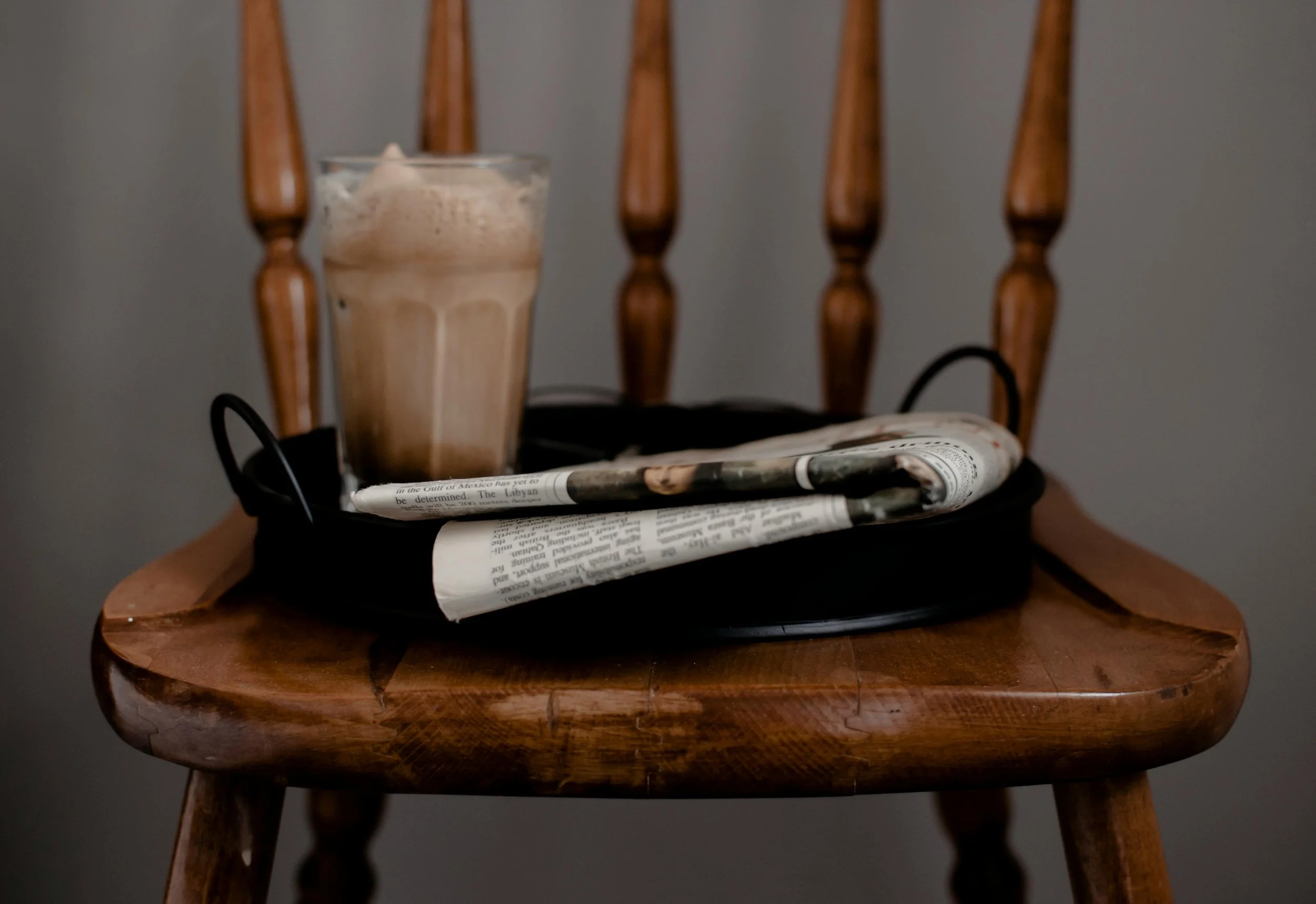 A glass of iced coffee with foam on top placed on a wooden chair with newspapers and black cords underneath.