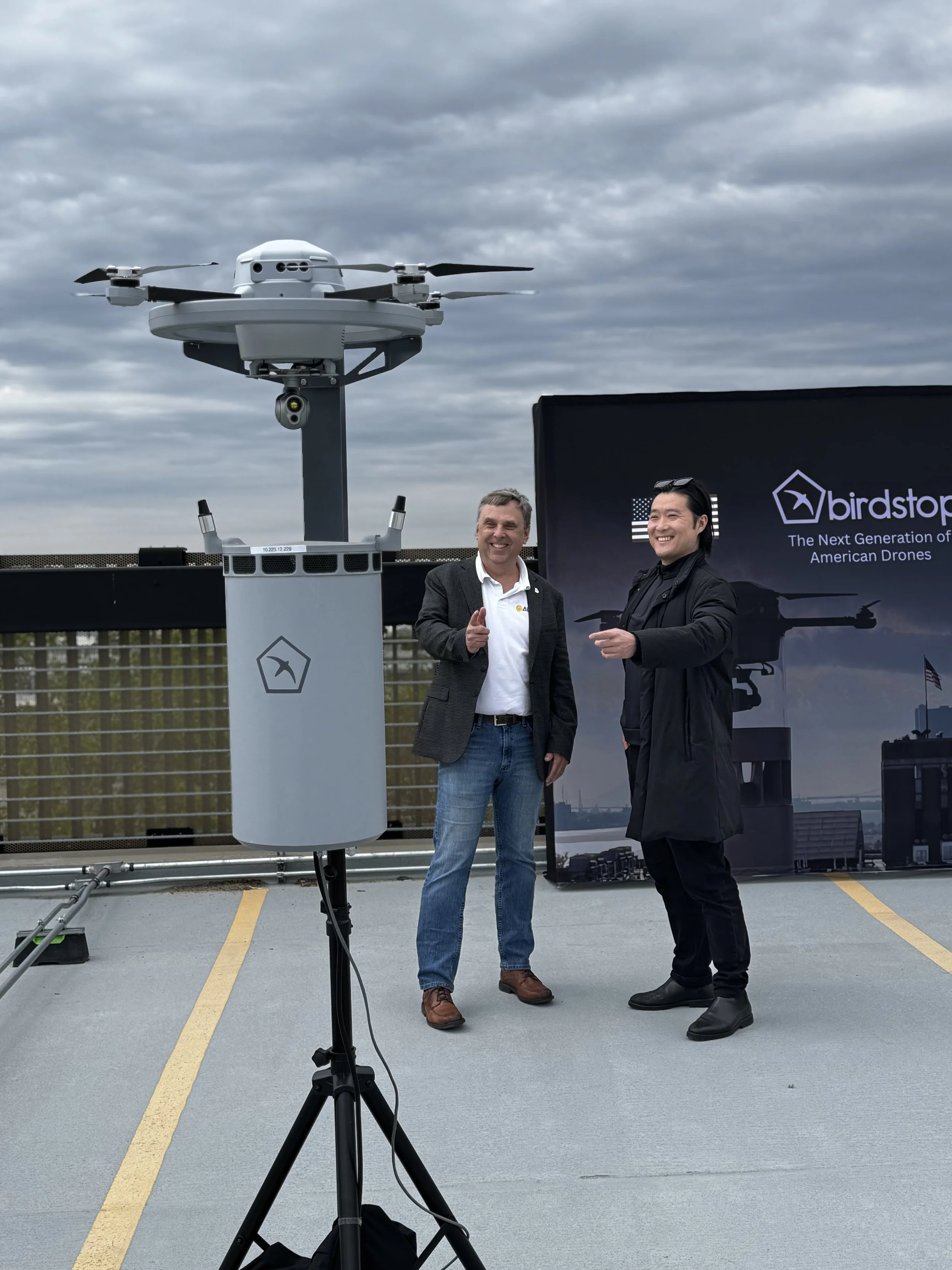 Two men standing on a rooftop, smiling and giving a thumbs up, with a drone on a tripod in the foreground and a sign that reads 'birdstop: The Next Generation of American Drones' in the background.