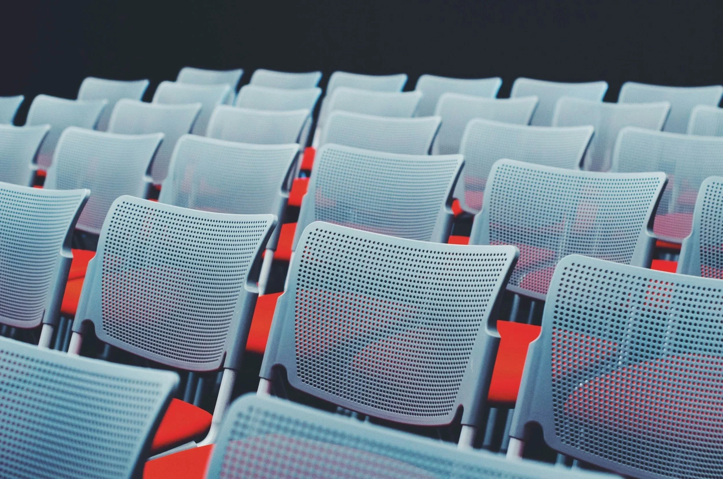 Multiple rows of light gray plastic chairs with black frames and red accents, arranged in a theater or conference room setting.