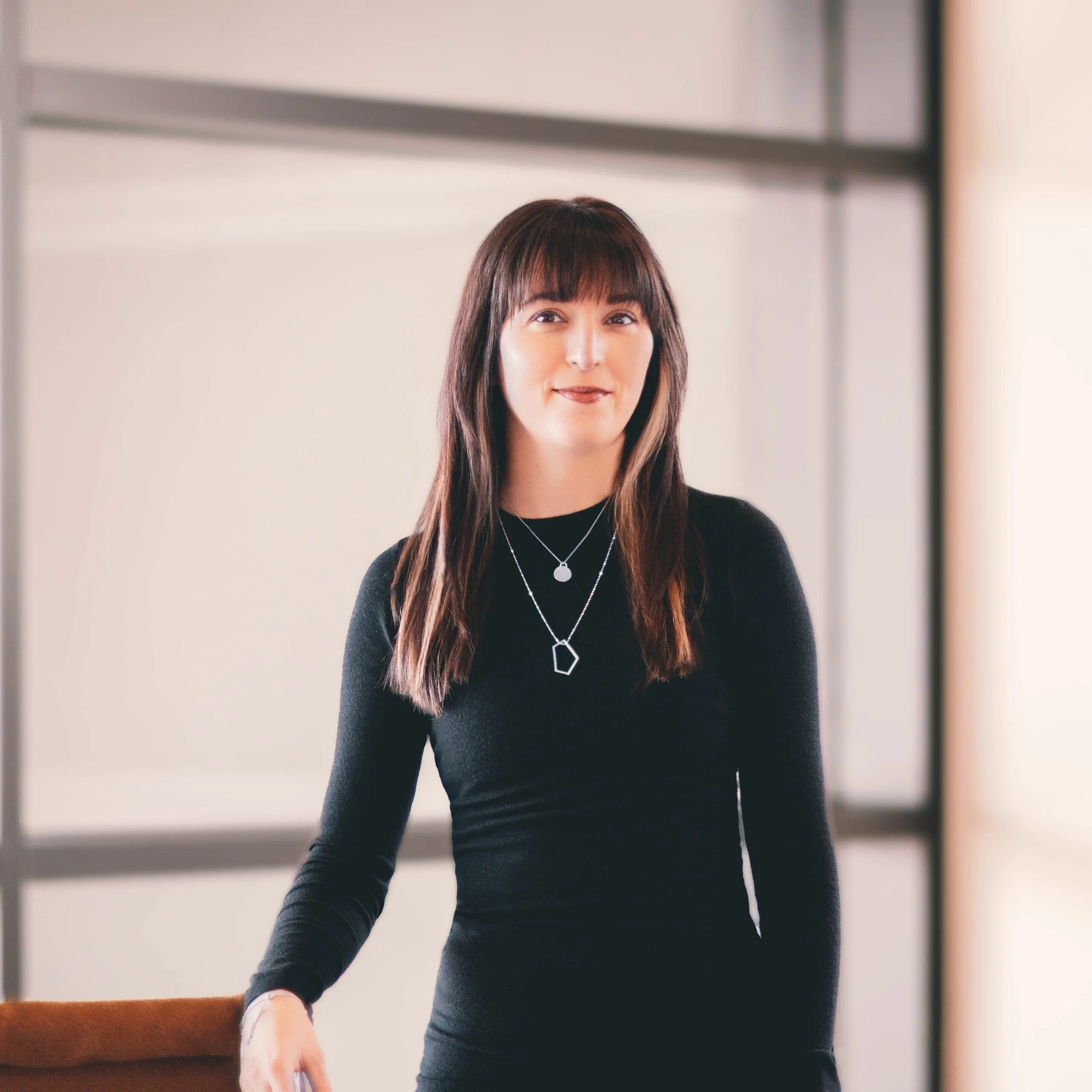 Close-up of a woman with long brown hair and bangs, wearing a black top and layered necklaces, standing indoors in front of a plain wall.