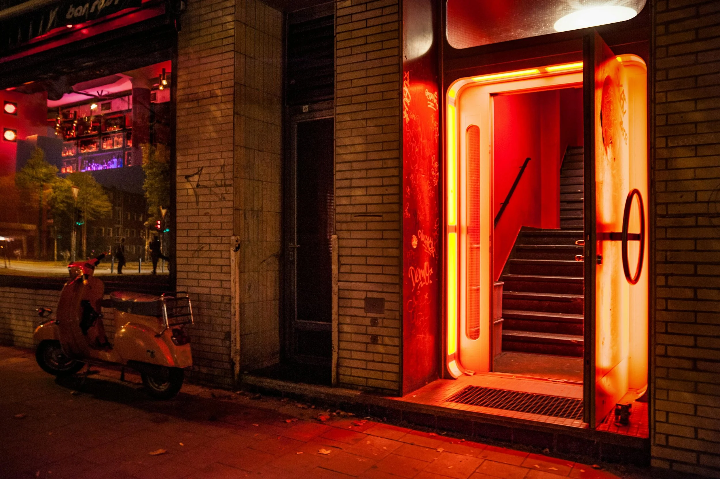 A neon-lit entrance to a building at night, with stairs leading up inside, flanked by brick walls. A scooter is parked outside, and a few people are walking on the street in the background.