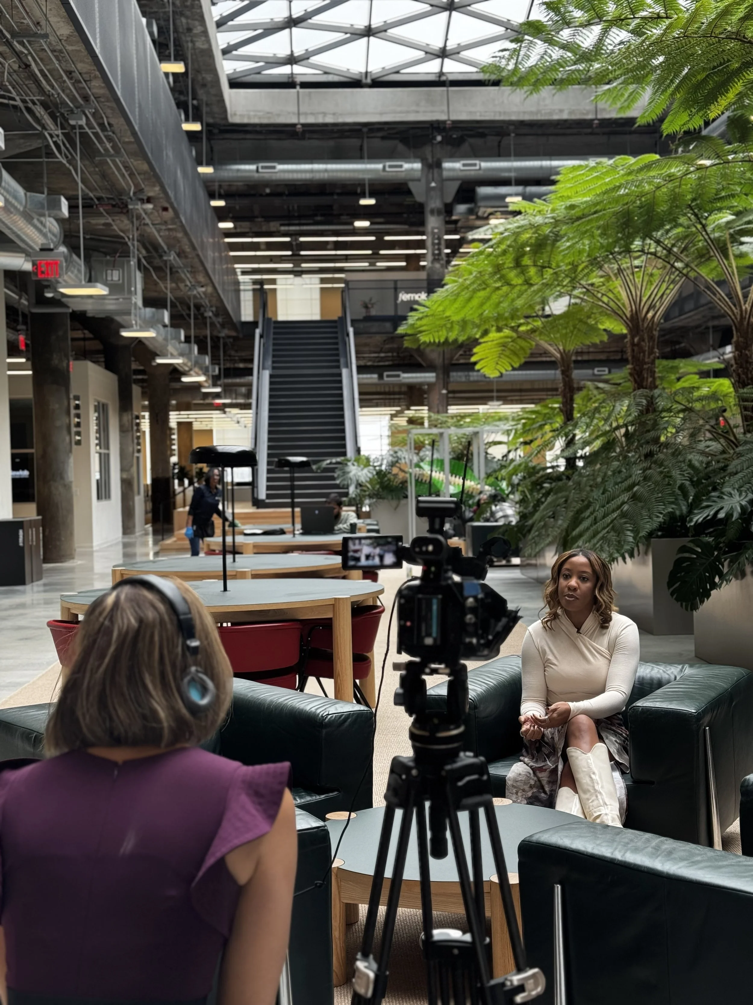 A woman sitting on a black leather couch being filmed by a camera operator in an industrial-style indoor setting with large green plants and a staircase in the background.