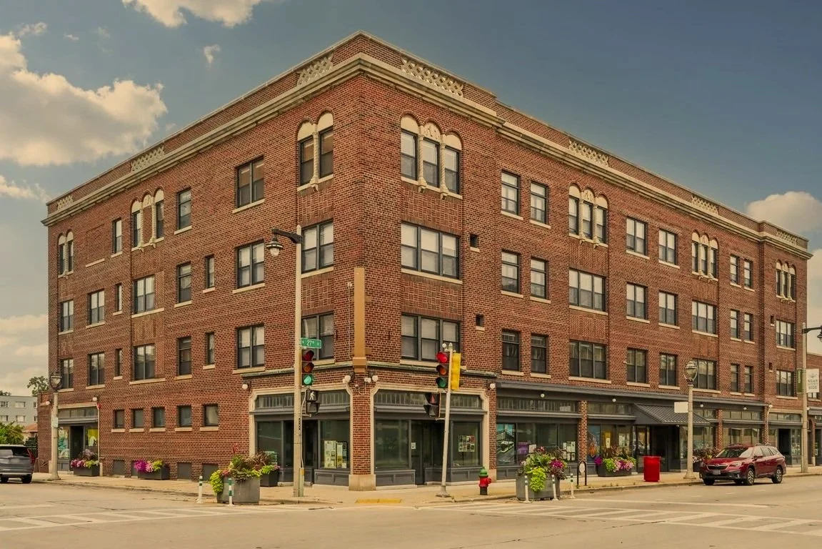 A four-story brick building with retail shops on the ground floor at a street corner under a partly cloudy sky.