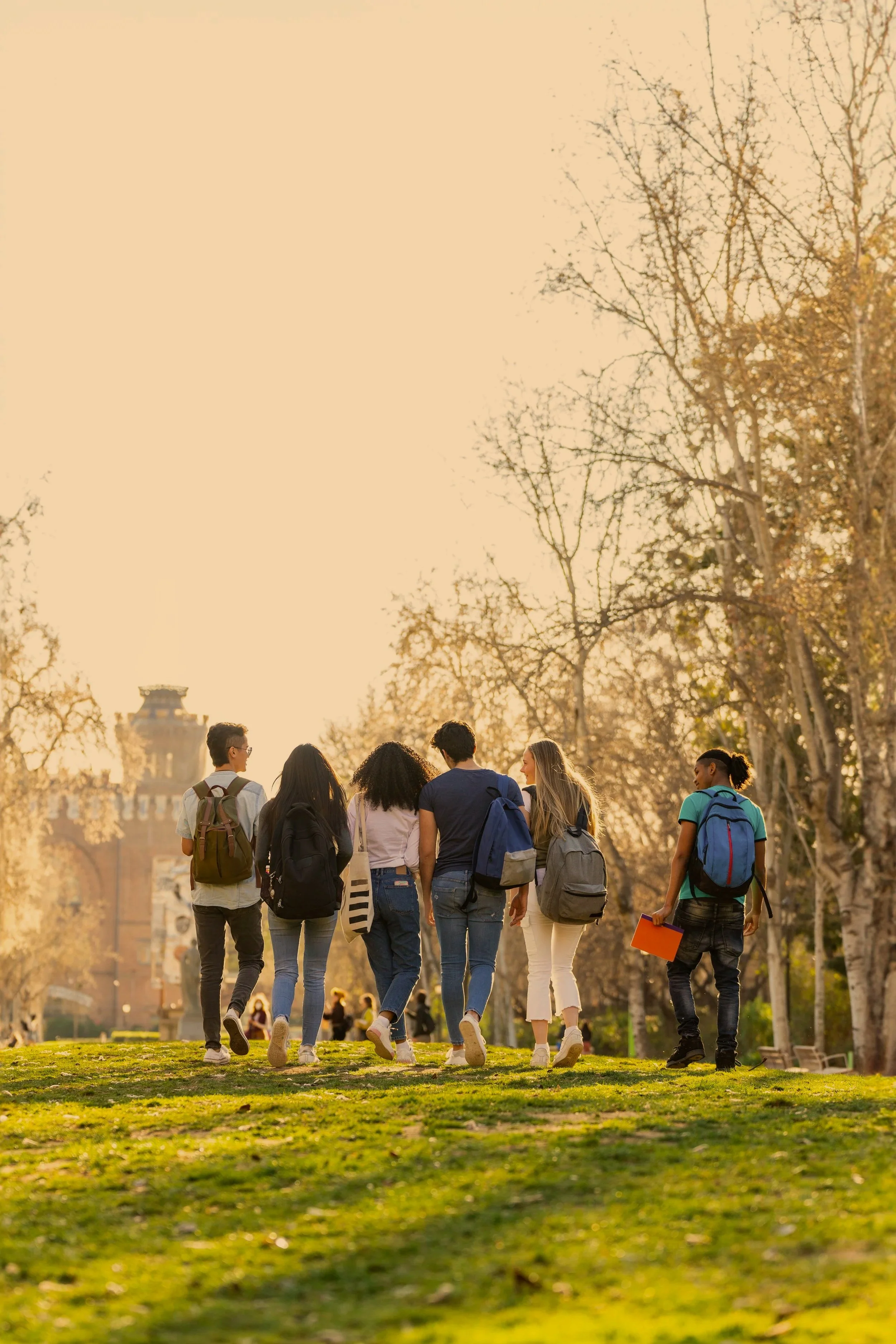 A group of diverse young people walking together in a park during sunset, carrying backpacks and books.