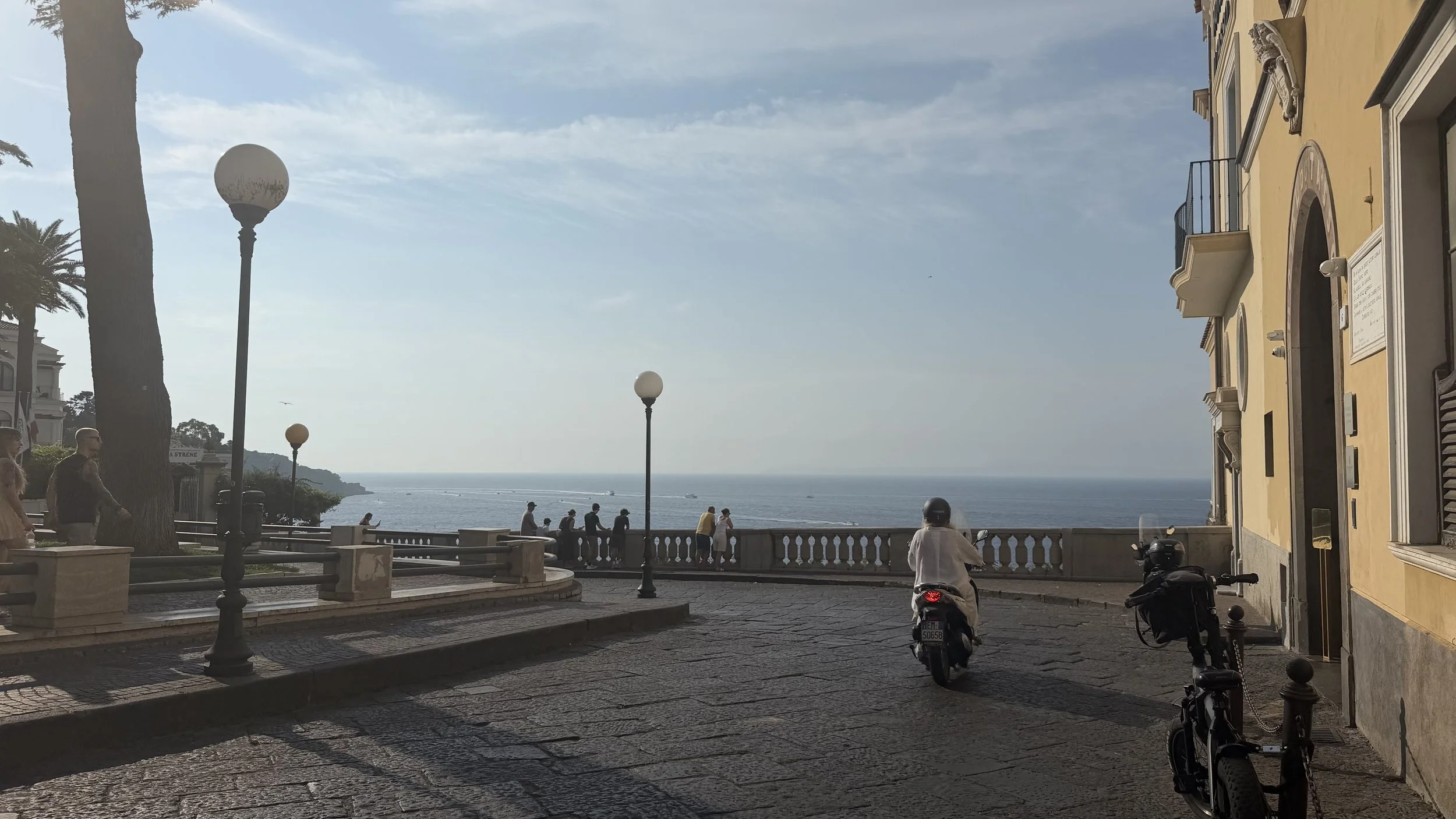 Seaside promenade with globe streetlamps, scooters, historic buildings, and ocean views