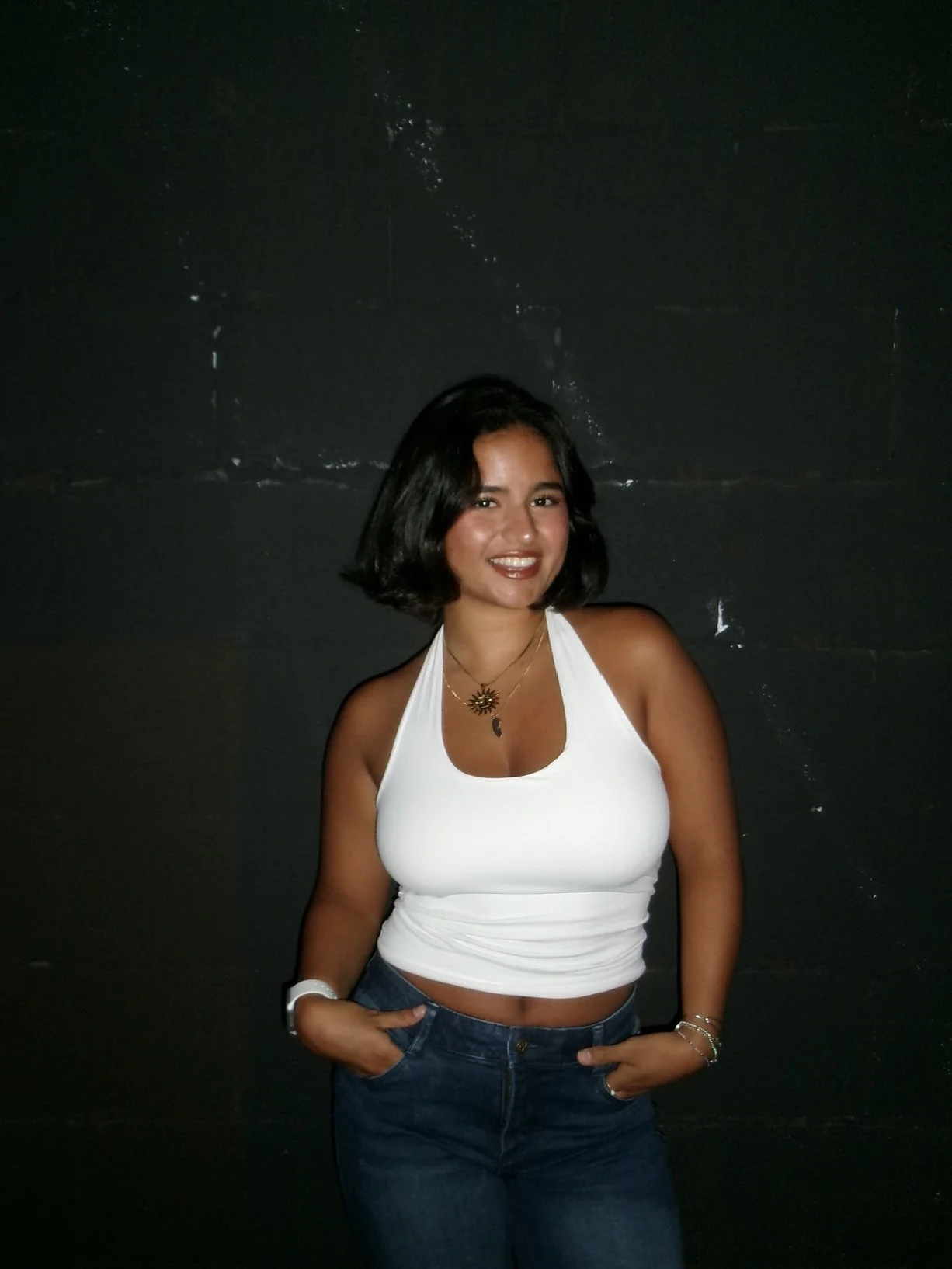 Smiling woman with short black hair wearing a white halter top, jeans, and layered necklaces against a dark wall