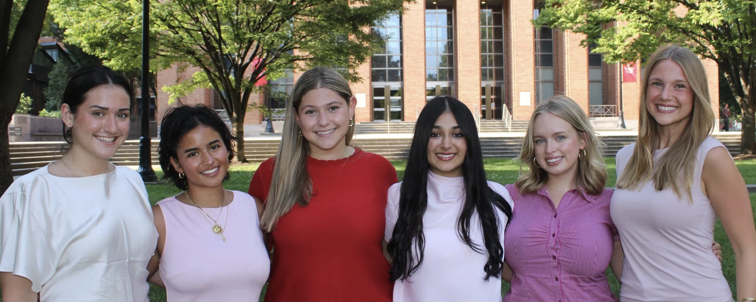 Six smiling women pose together on a leafy college campus in front of a brick building.