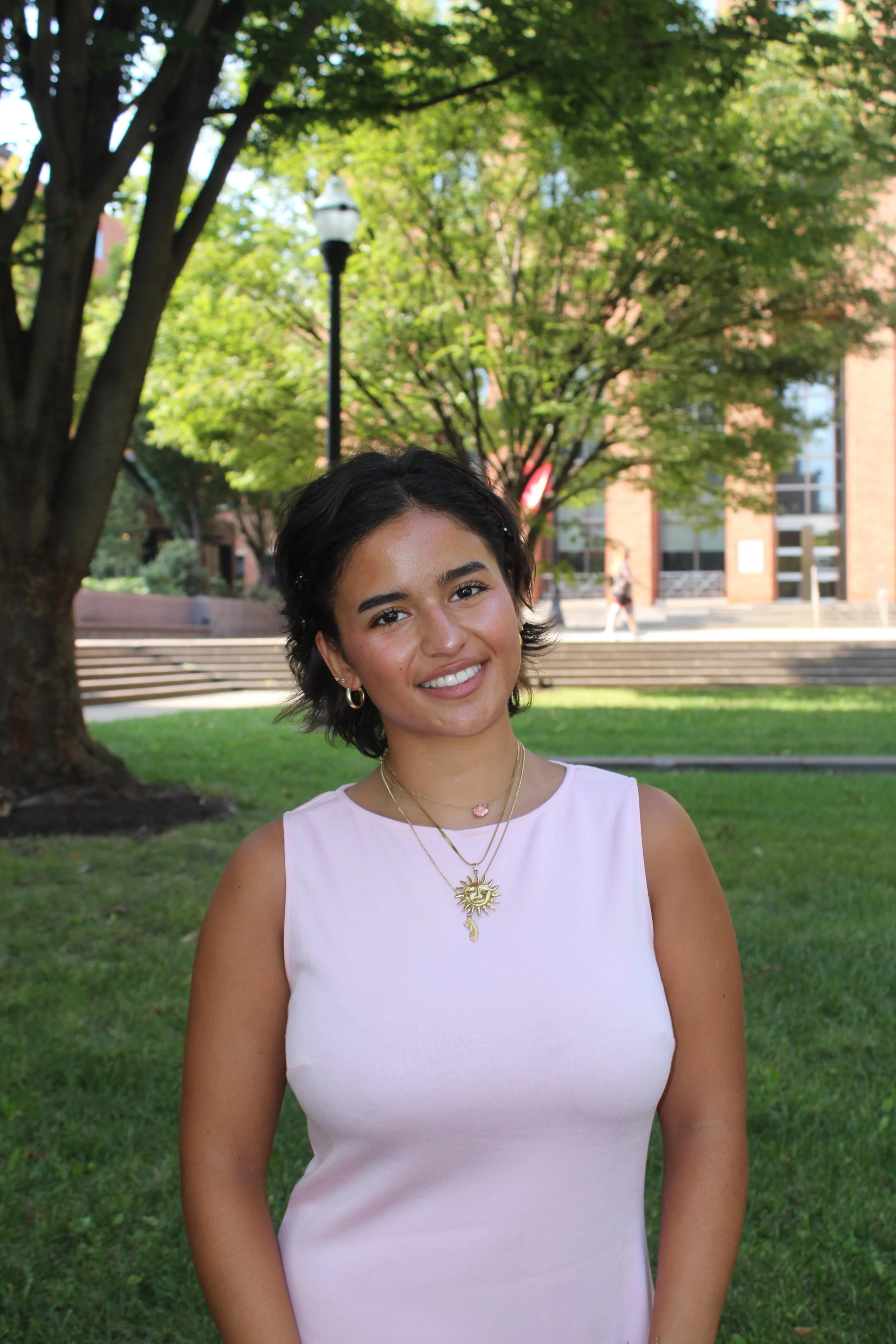 Smiling woman in a pink sleeveless top and layered necklaces standing outdoors on a tree-lined campus.