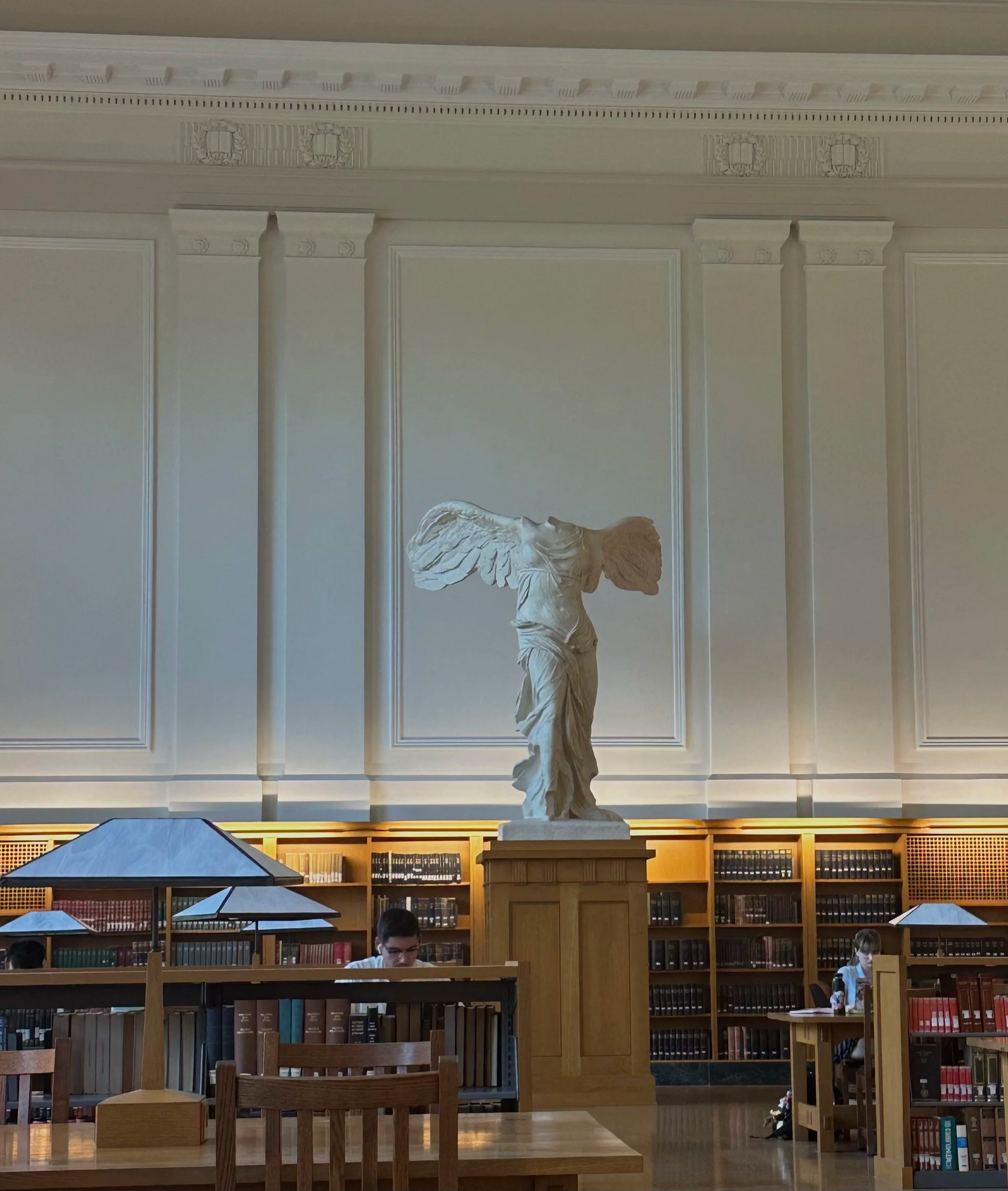 Winged Victory of Samothrace replica displayed in an ornate library reading room with bookshelves and study tables