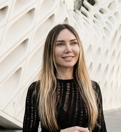 Close-up of a blonde female wearing a black sweater, smiling, standing in front of the Broad Museum in Los Angeles.