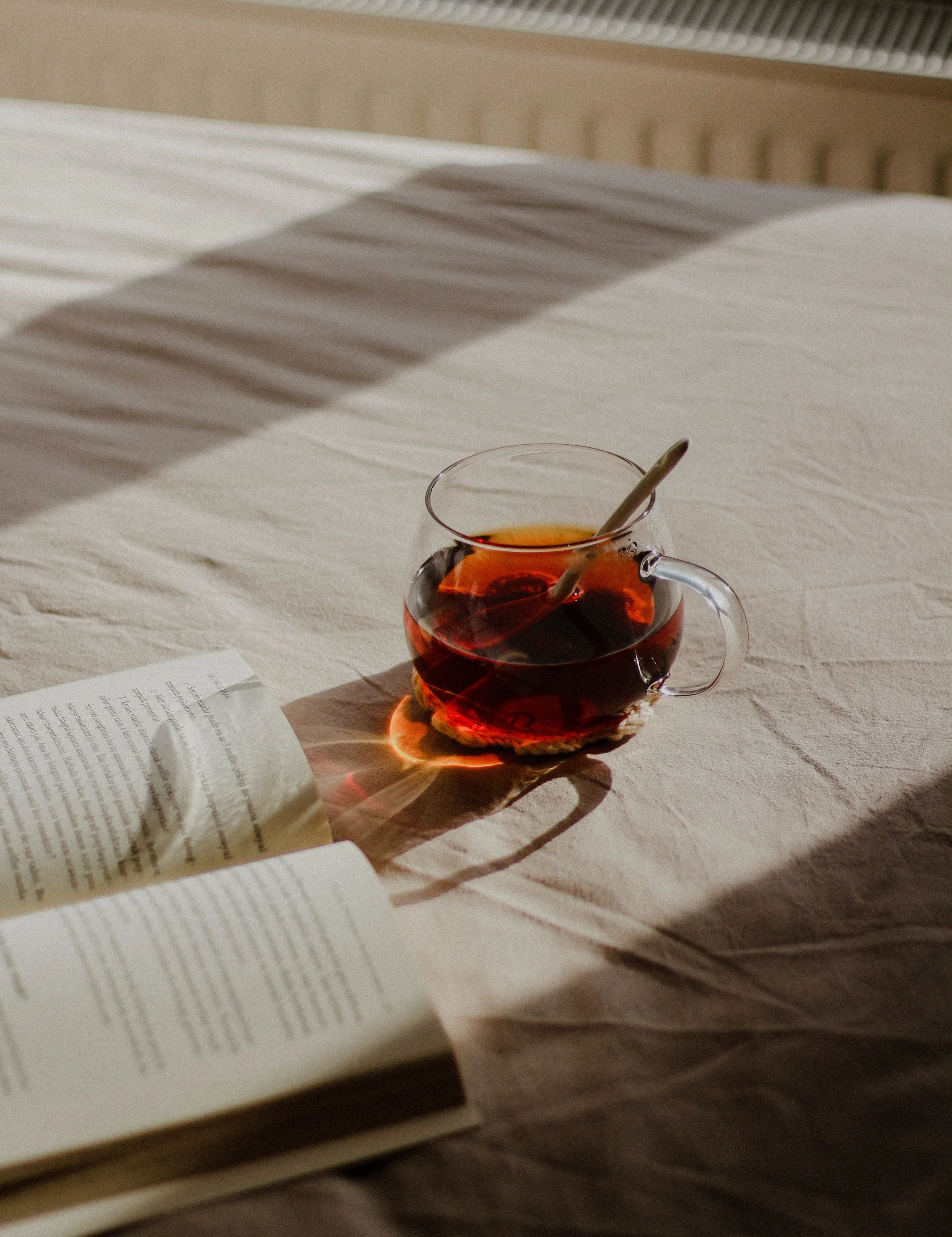 A glass cup filled with tea, placed on a cloth coaster on a beige bedspread. There is an open book partially visible in the foreground, and sunlight casts shadows across the scene.