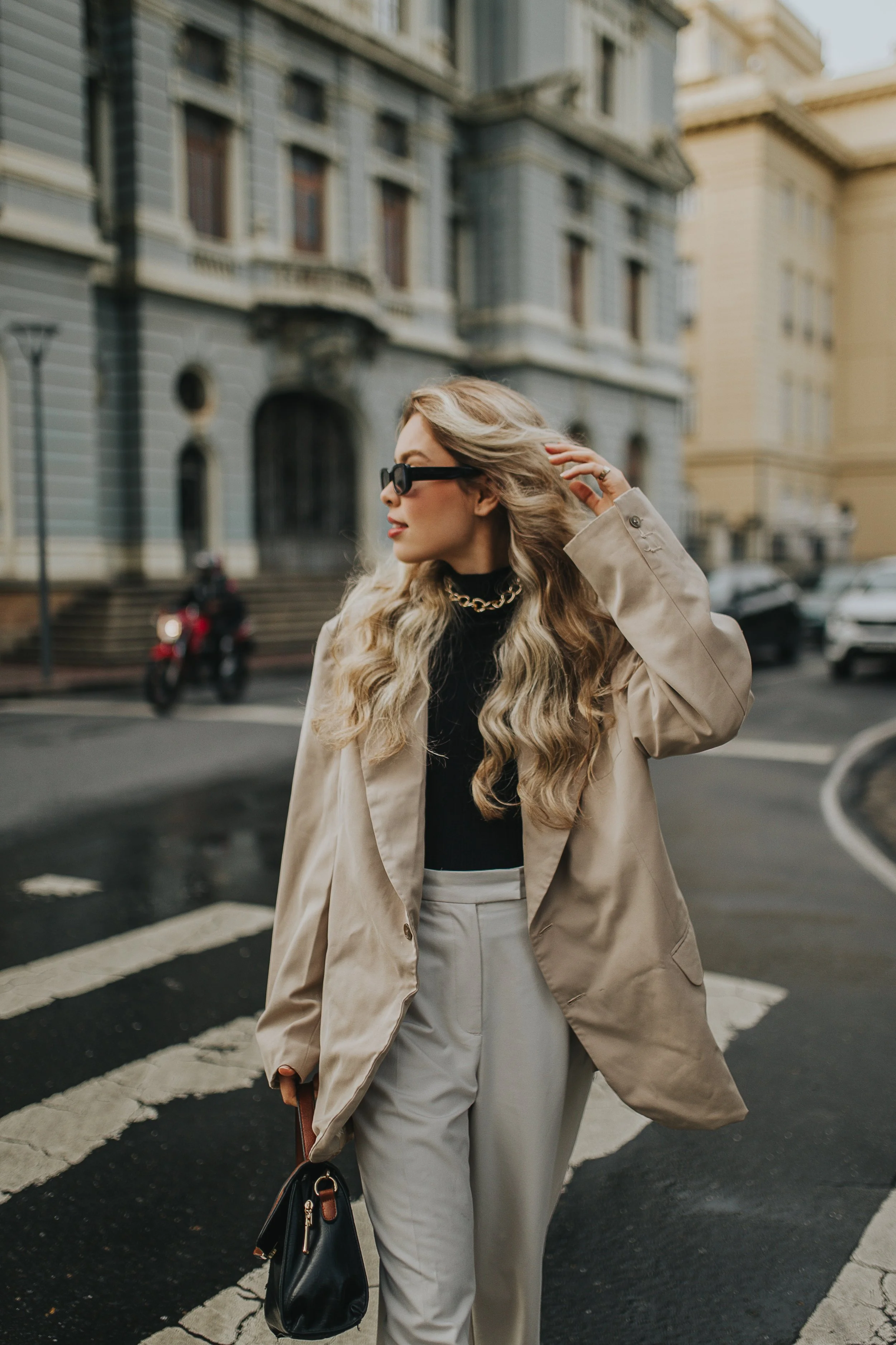 A woman with long blonde hair wearing black sunglasses, a beige blazer, black top, and beige trousers, walking across a city street with historic buildings in the background, carrying a black handbag.