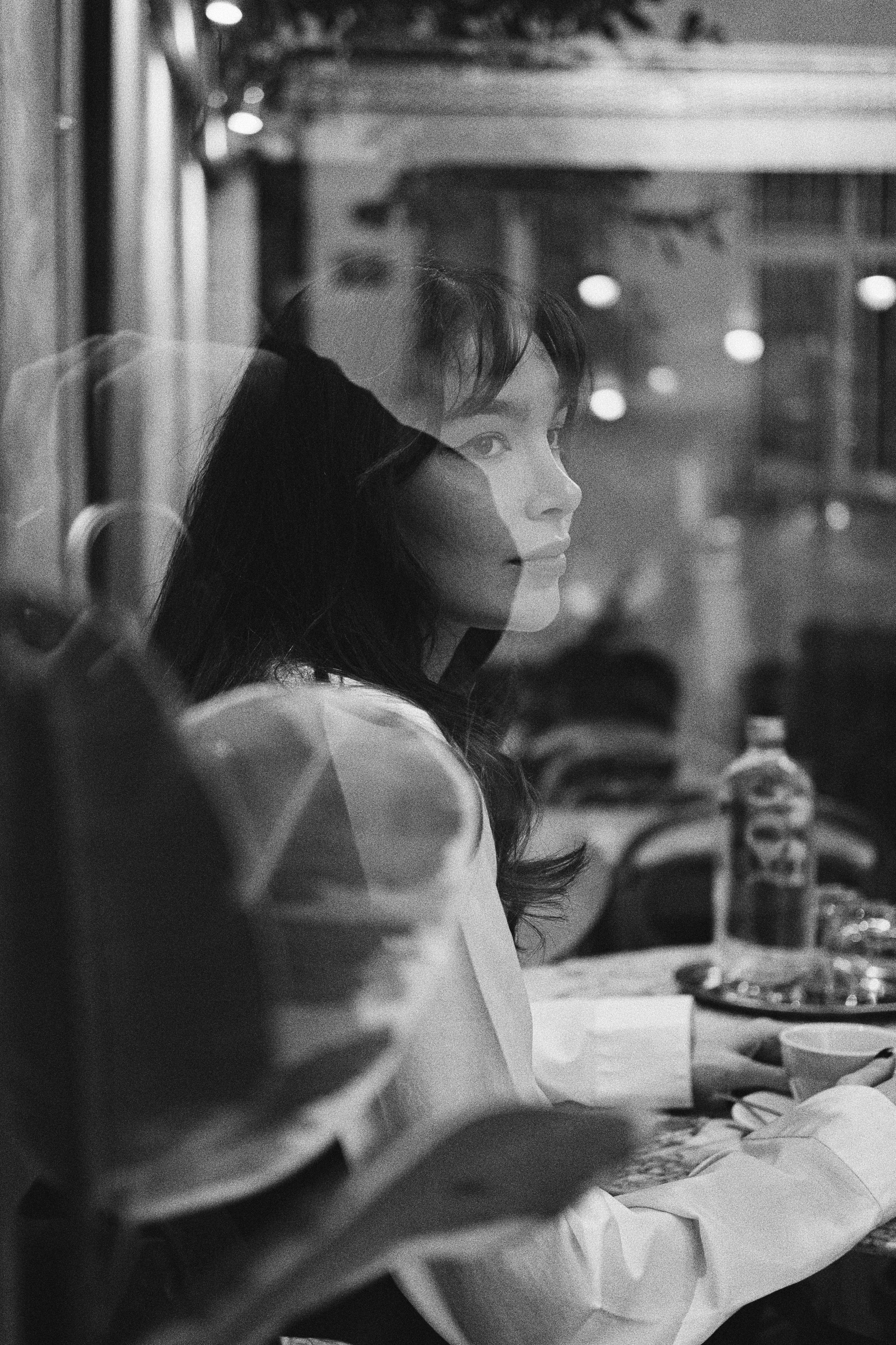 A woman with long dark hair and light skin sitting at a table in a restaurant or cafe, looking to the side with a calm expression, surrounded by bottles and plates on the table, with decorative lights in the background.