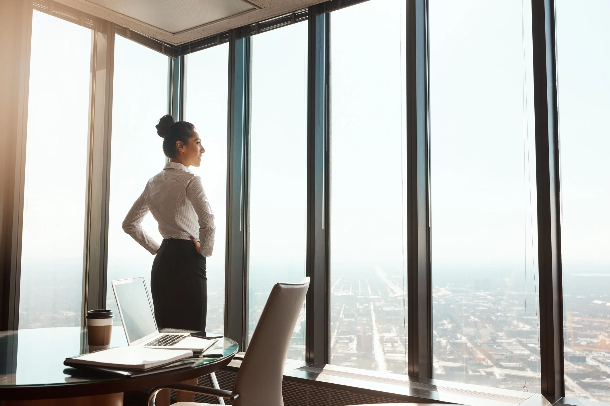 Young successful brunette female looking out of windows in office, laptop open on desk.