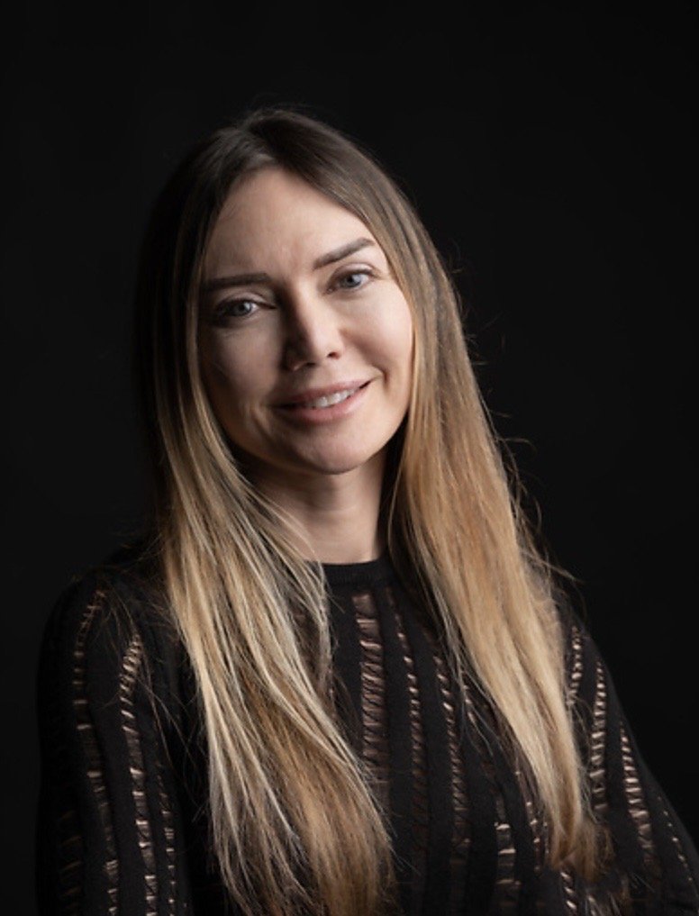 A woman with long, light brown hair and fair skin, smiling and wearing a black top with cut-out patterns, against a black background.