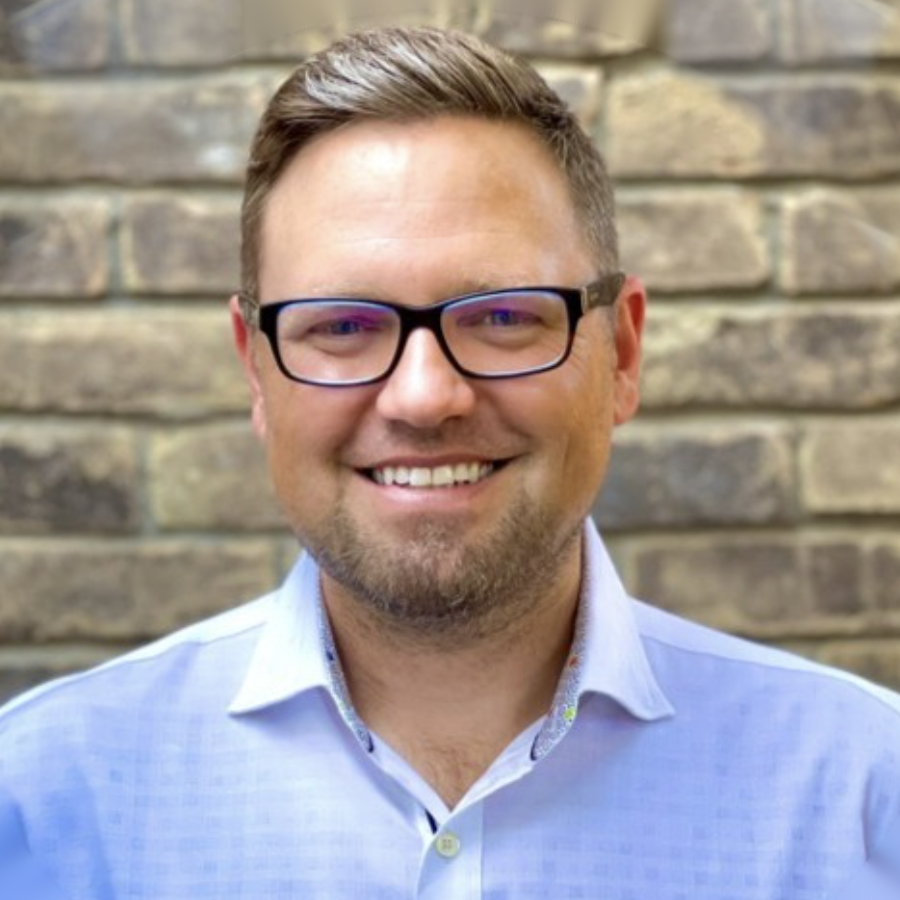 Blonde man with glasses smiling in front of brick wall