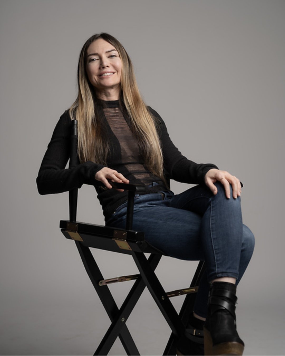Woman with long hair sitting on a director's chair, smiling and looking at the camera, wearing a black top and jeans, against a plain gray background.