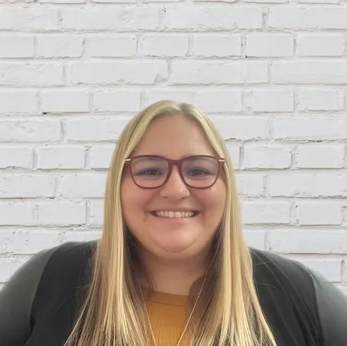 A smiling woman with long blonde hair wearing glasses, a black jacket, and a mustard-colored top, sitting in front of a white brick wall.