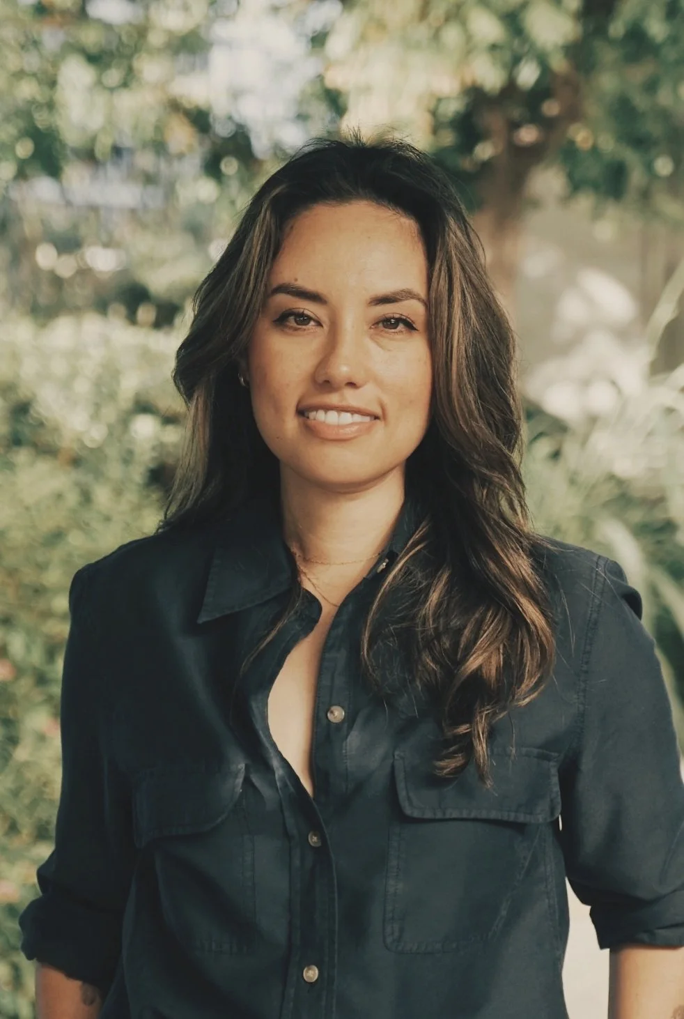 A woman with long brown hair wearing a dark button-up shirt, smiling outdoors with trees and greenery in the background.