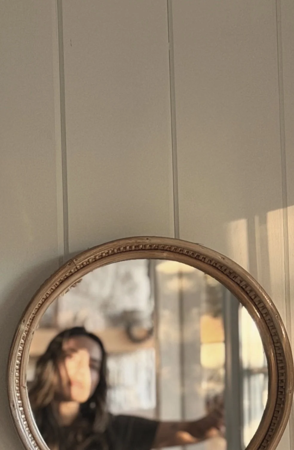 Close-up of a decorative oval mirror with a beaded frame, hanging on a light-colored wall. The reflection shows a woman with long hair taking a selfie inside a room with large windows and some blurred decorative items.
