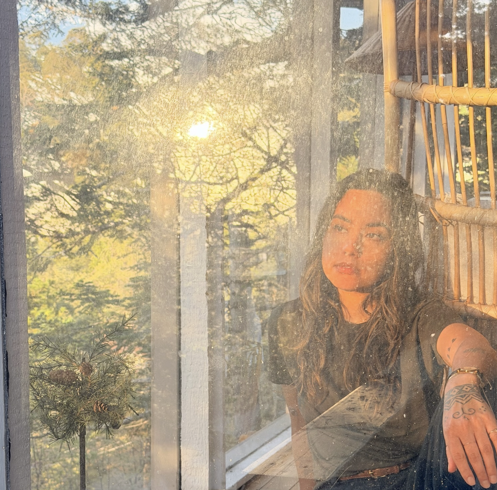 A woman with long, wavy hair sitting near a window with sunlight shining through, and a view of trees outside. She has tattoos on her left arm and appears relaxed.