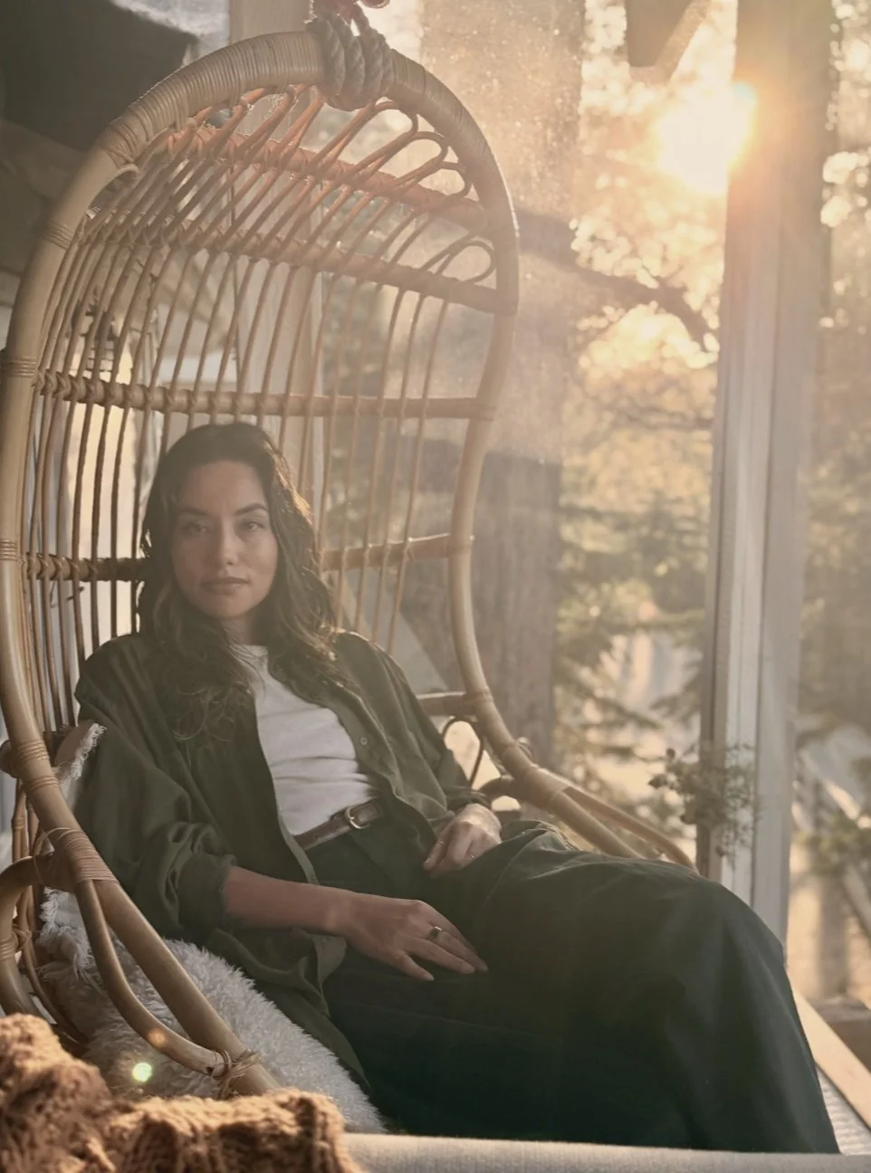 Woman relaxing in a wicker hanging chair on a balcony, sun setting behind trees, warm golden light.