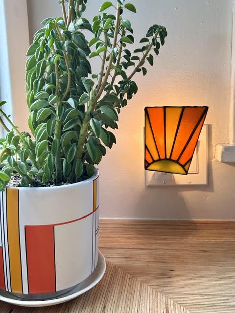 Indoor plant in a decorative pot on a wooden surface next to a wall mounted lamp with a geometric orange and yellow design