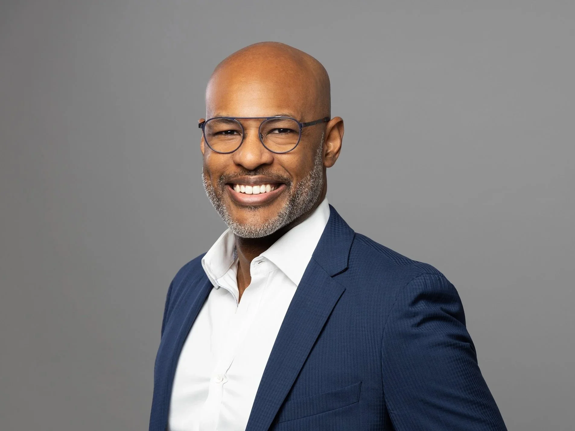 Smiling middle-aged man with glasses, bald head, and gray beard, wearing a navy blue suit and white shirt, against a gray background.