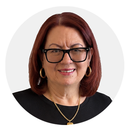 A middle-aged woman with red hair, black glasses, wearing gold hoop earrings and a gold necklace, smiling slightly with a concerned expression against a plain background.