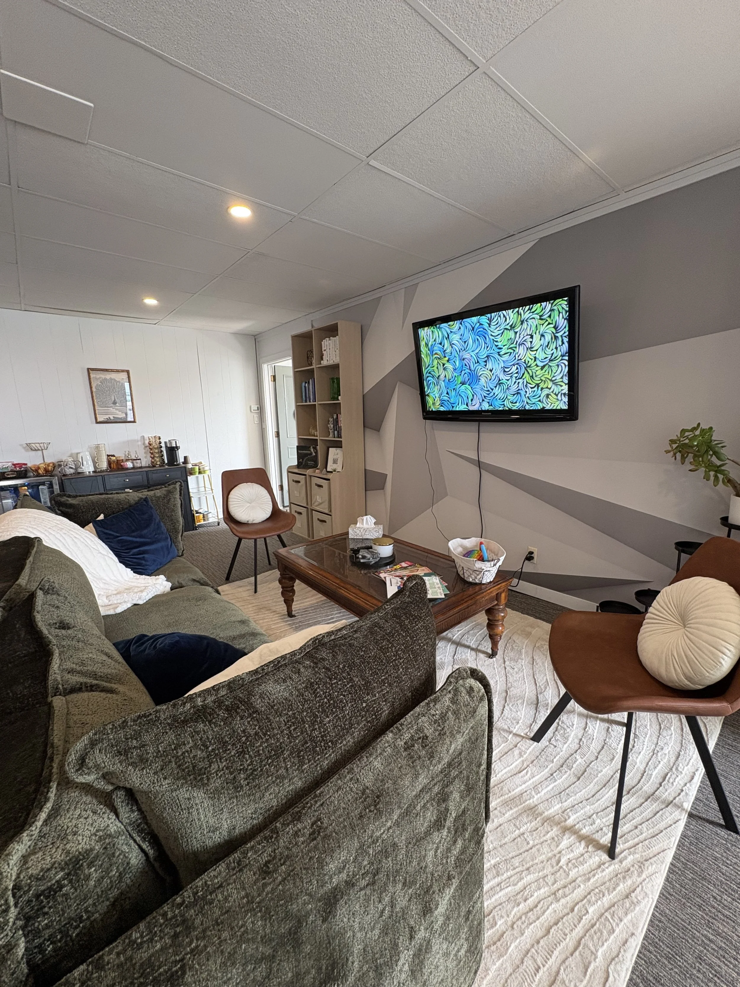Living room with gray walls, a dark gray patterned sofa, a wooden coffee table with various items, a white bookshelf filled with books and decor, a brown chair with a white round pillow, a TV mounted on the wall displaying a colorful abstract pattern, a print on the wall, and a rug on the floor.