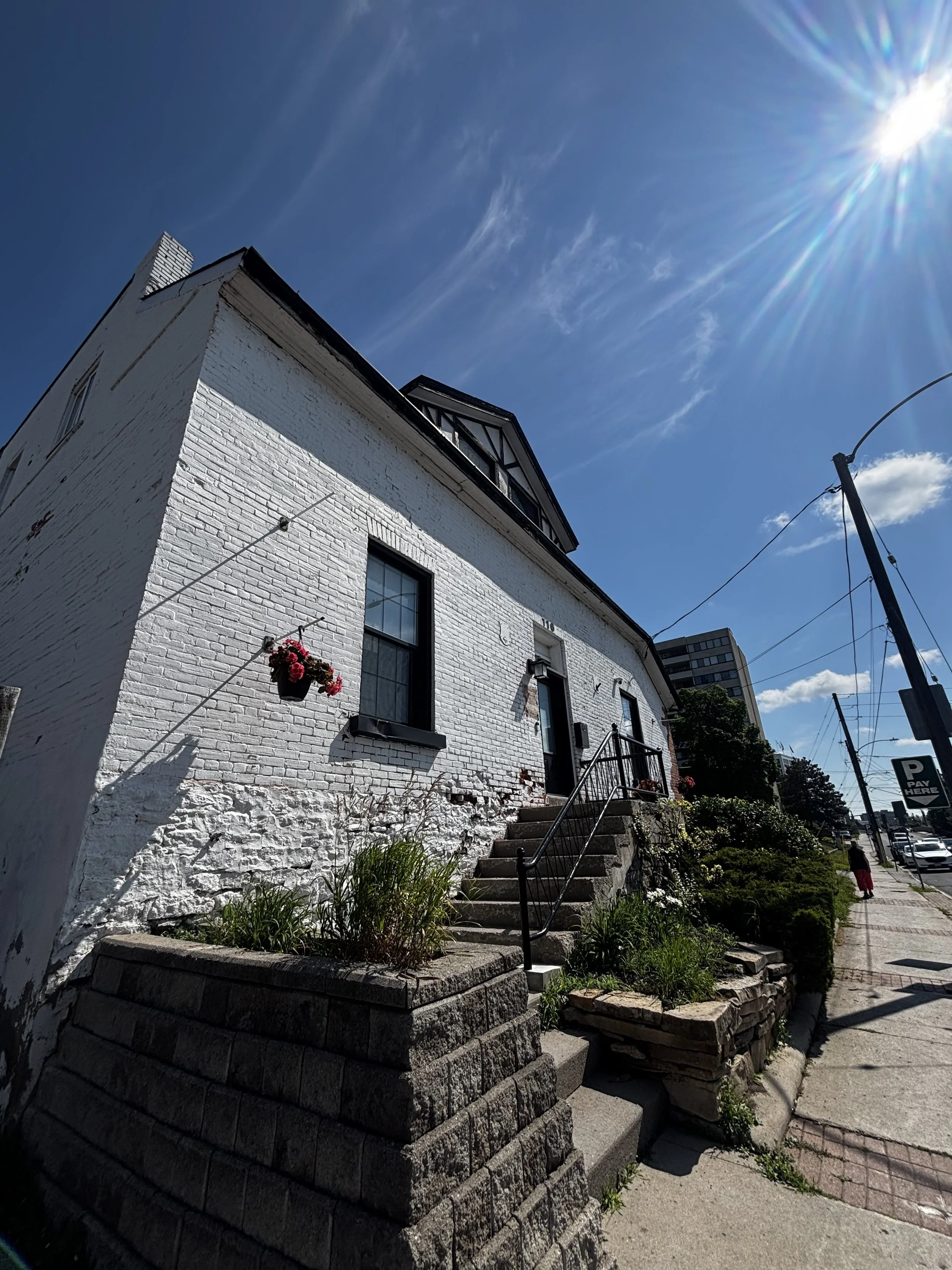 Horizon Wellness Collective is in a White brick building with black windows and a black roof under a bright blue sky with sun glare, street, sidewalk, and plants in the foreground in Peterborough Ontario.