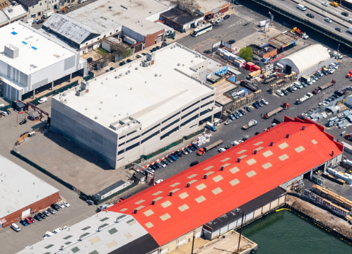 Aerial view of a commercial area with a large white multi-story building, a warehouse with a red roof, and surrounding parking lots filled with cars.