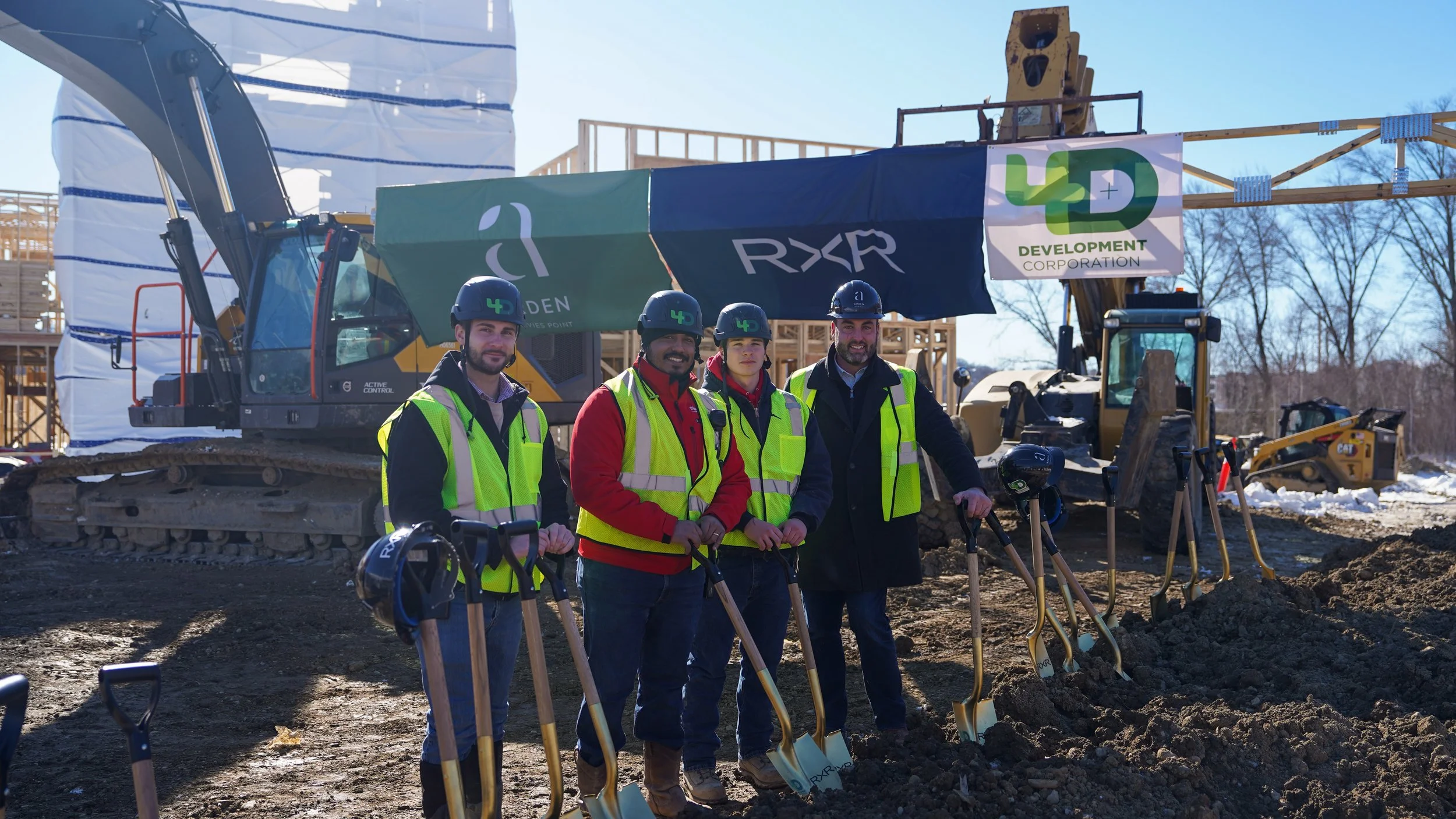Four construction workers in safety vests and helmets standing in front of heavy equipment on a construction site, with a wooden building framework and trees in the background.