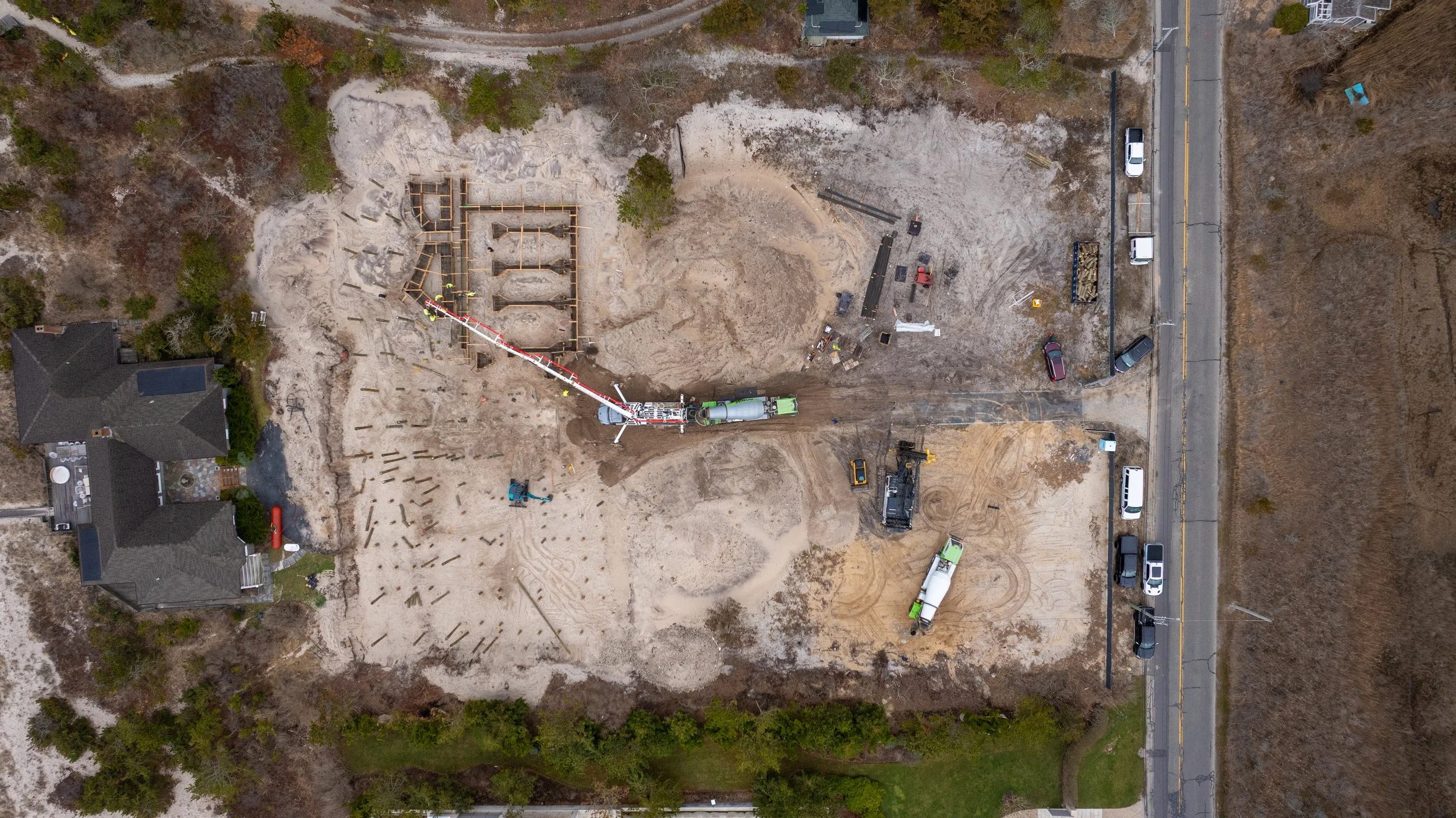 Aerial view of a construction site with a foundation being built, construction vehicles, workers, and surrounding houses and trees.