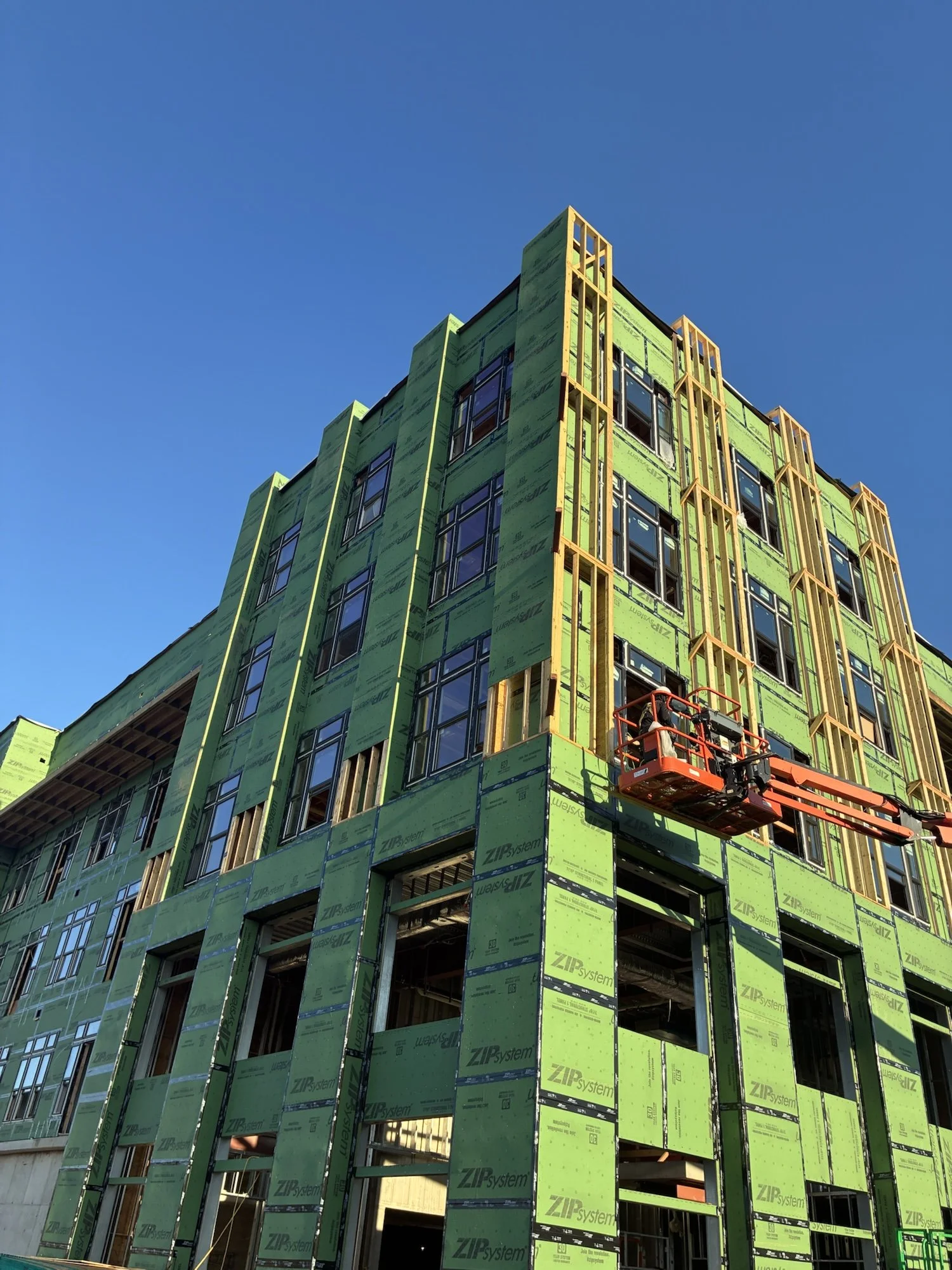 A multi-story building under construction with green insulation panels and large windows. A construction lift is visible on the lower right side, and the sky is clear and blue.