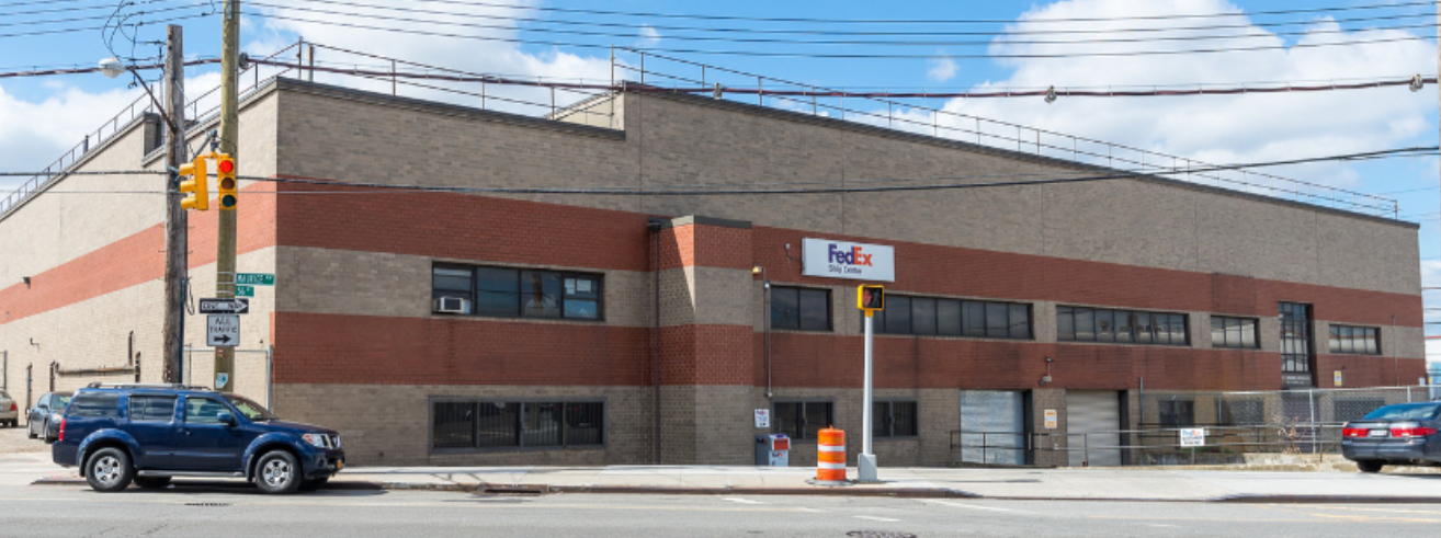 A FedEx shipping center building with a sidewalk in front, cars parked along the street, traffic lights, and overhead power lines. The building has a brick facade with a FedEx logo sign.