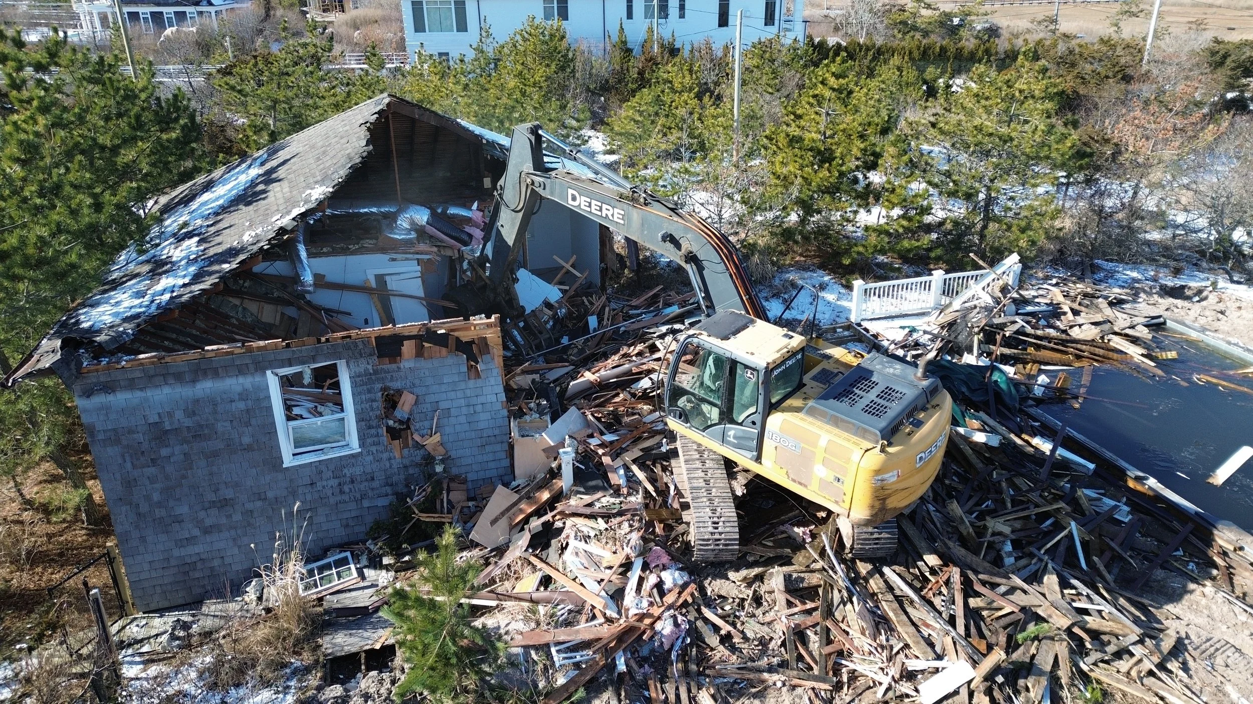 An aerial view of a house being demolished with a bulldozer, surrounded by trees, snow, and debris.