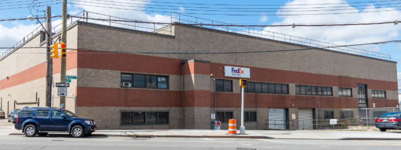 A large commercial building with a FedEx sign, multiple windows, and a garage door. There are cars parked in front and a traffic light at the intersection.