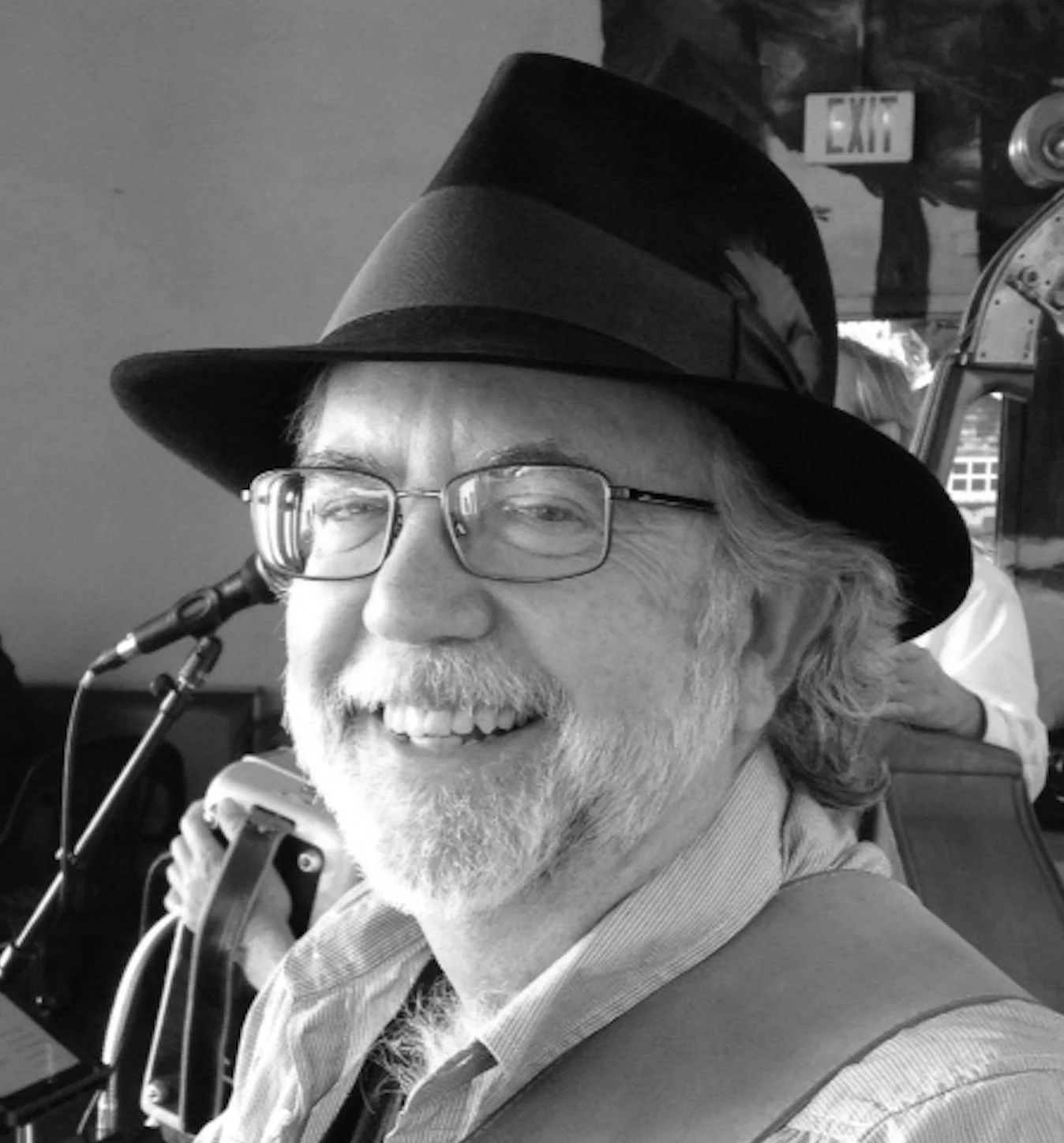 A smiling older man with glasses and a beard wearing a black fedora hat. He's holding a microphone and appears to be in a social setting, with a person and an exit sign visible in the background.