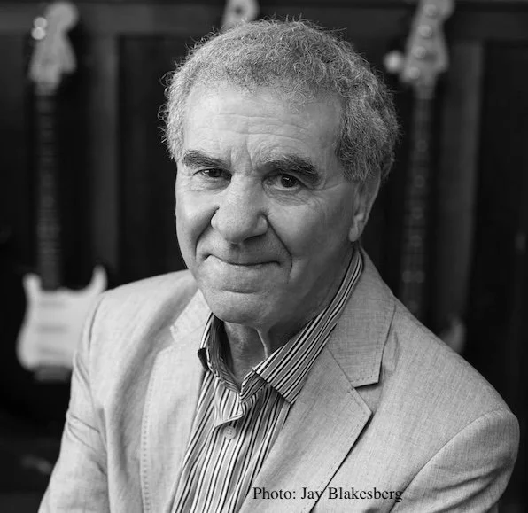 Black and white portrait of an older man wearing a blazer and striped shirt, with a background featuring guitars and musical equipment.