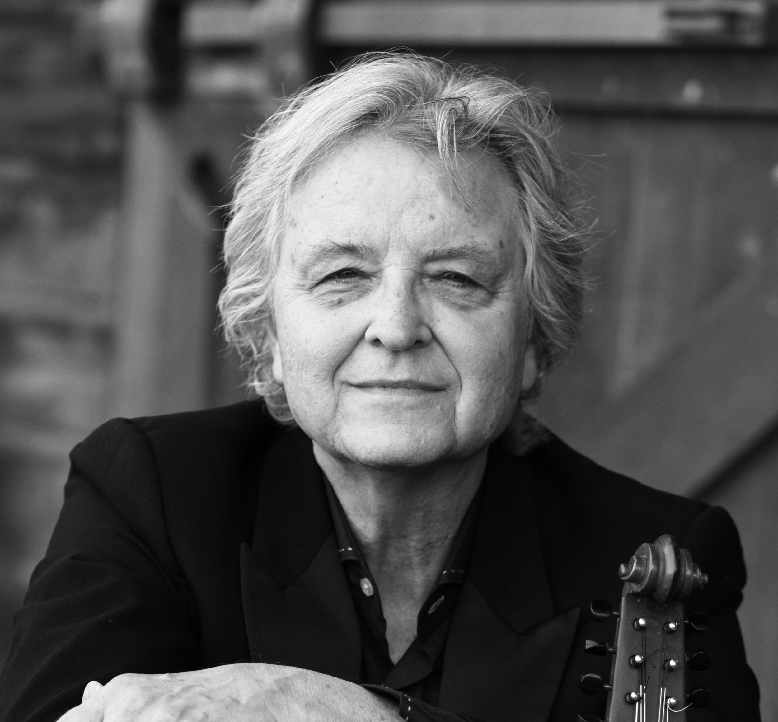 Black and white close-up photo of an older woman with short, wavy hair, smiling, wearing a dark top, seated with a guitar.