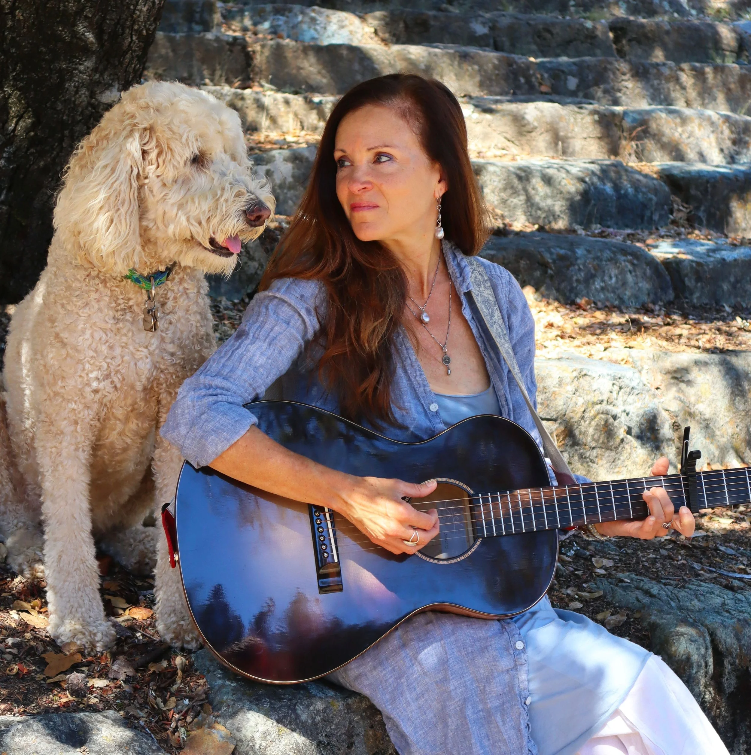 Woman with long reddish-brown hair sitting outdoors on rocks, playing an acoustic guitar, with a fluffy cream-colored dog standing nearby, in a wooded area with fallen leaves.