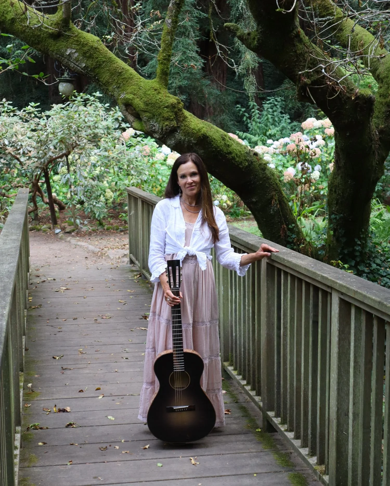 A woman with long brown hair wearing a white blouse and long skirt standing on a wooden bridge, holding an acoustic guitar, in a lush green forest with moss-covered tree branches and pink and white flowers in the background.