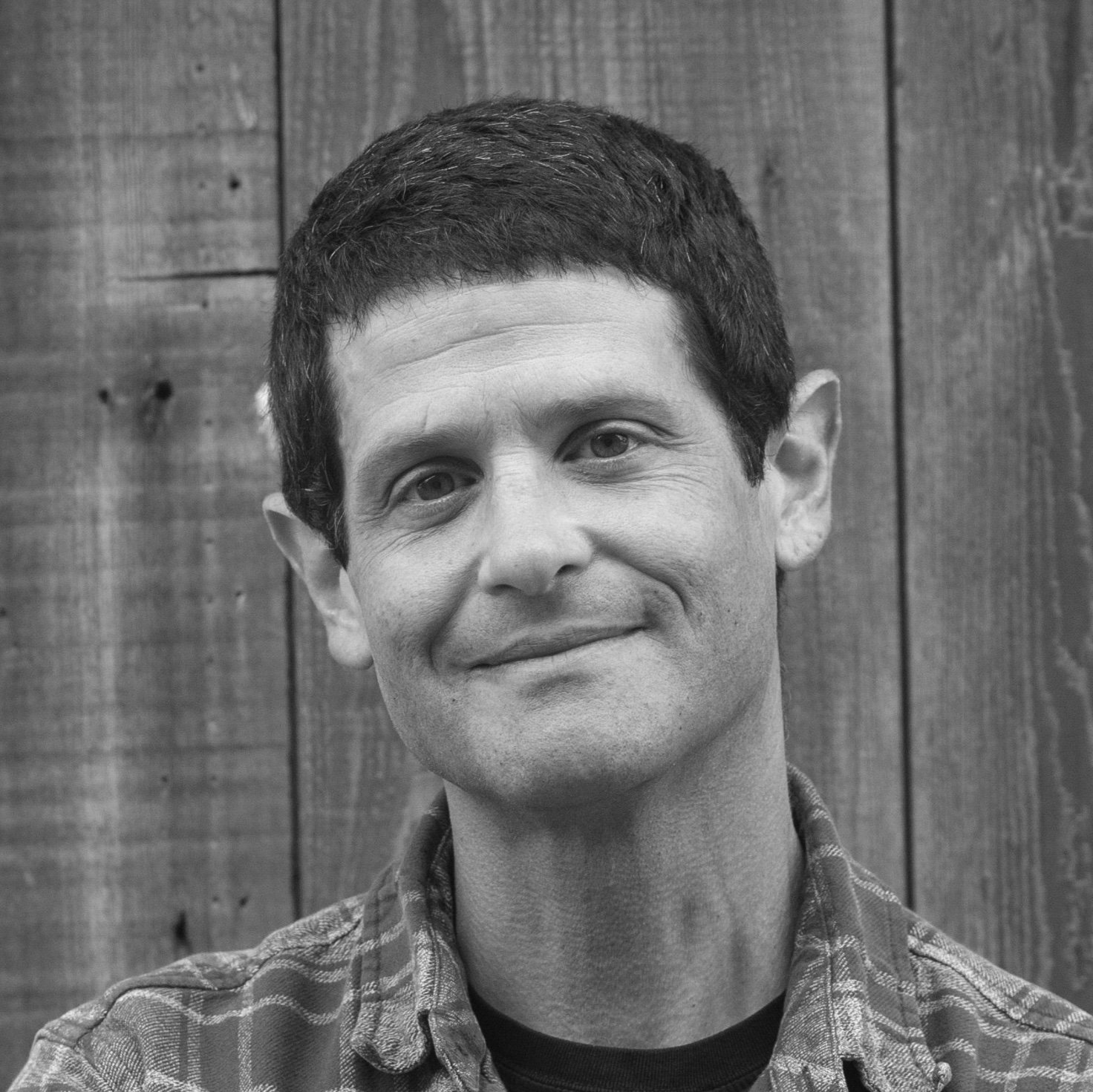 Black and white portrait of a man smiling, with a wooden wall in the background. He has short dark hair and is wearing a checkered shirt.