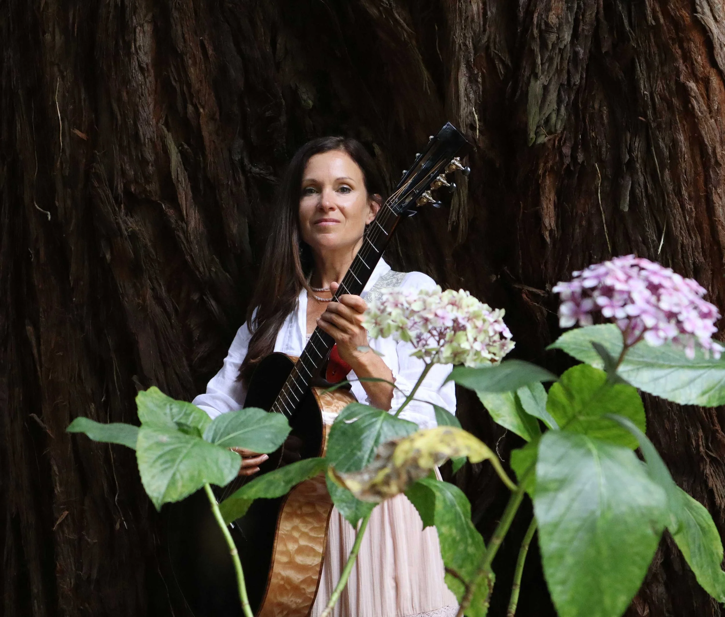 Woman with long dark hair holding a guitar in front of a large tree trunk, surrounded by green leaves and pink flowers.