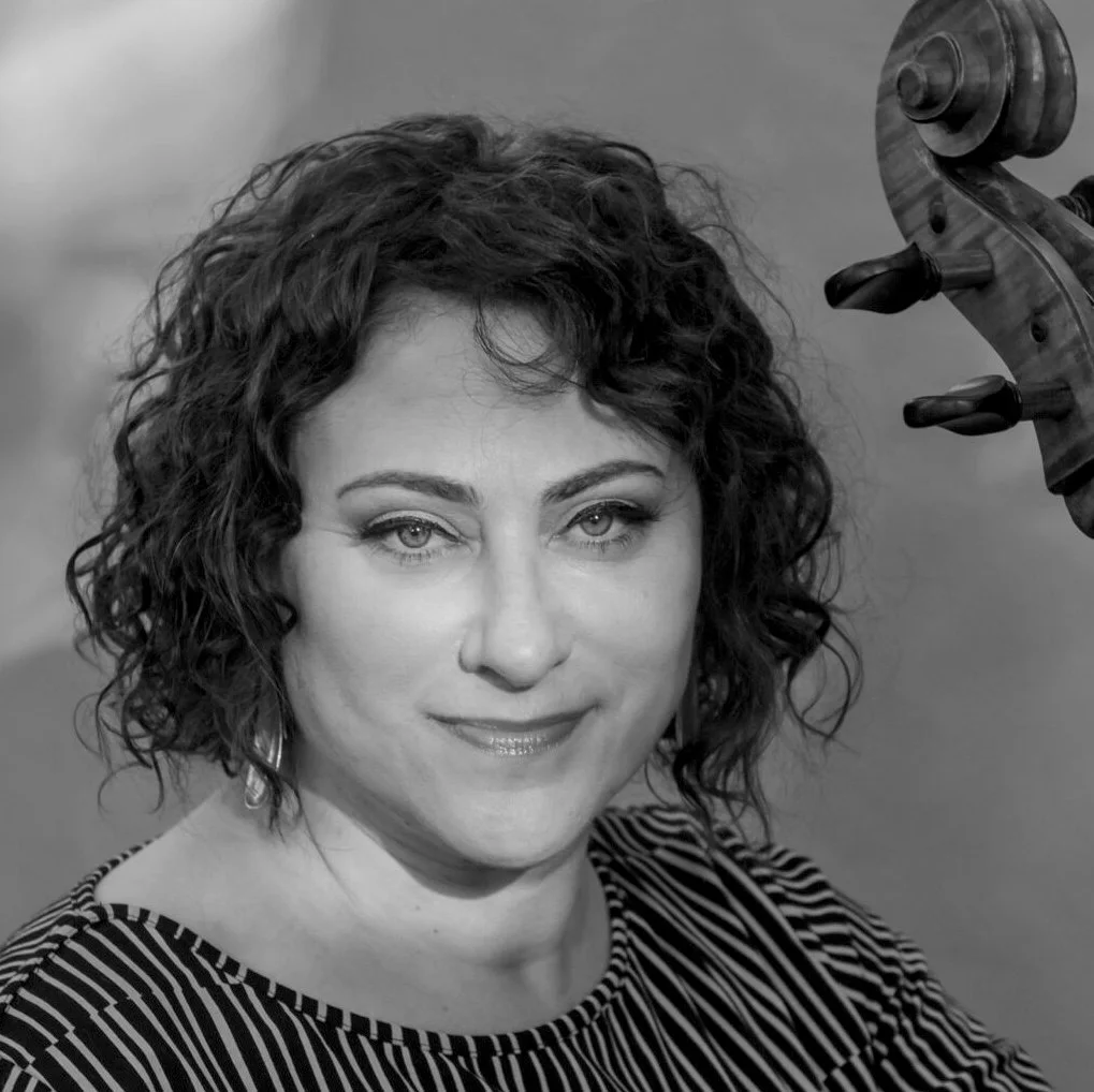 Black and white photo of a woman with short curly hair smiling at the camera, holding a violin bow.