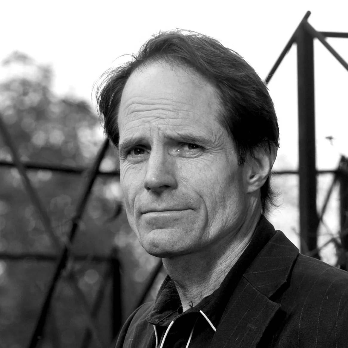 Close-up black and white portrait of a middle-aged man with dark hair, outdoors, with blurred trees and metal structure in background.