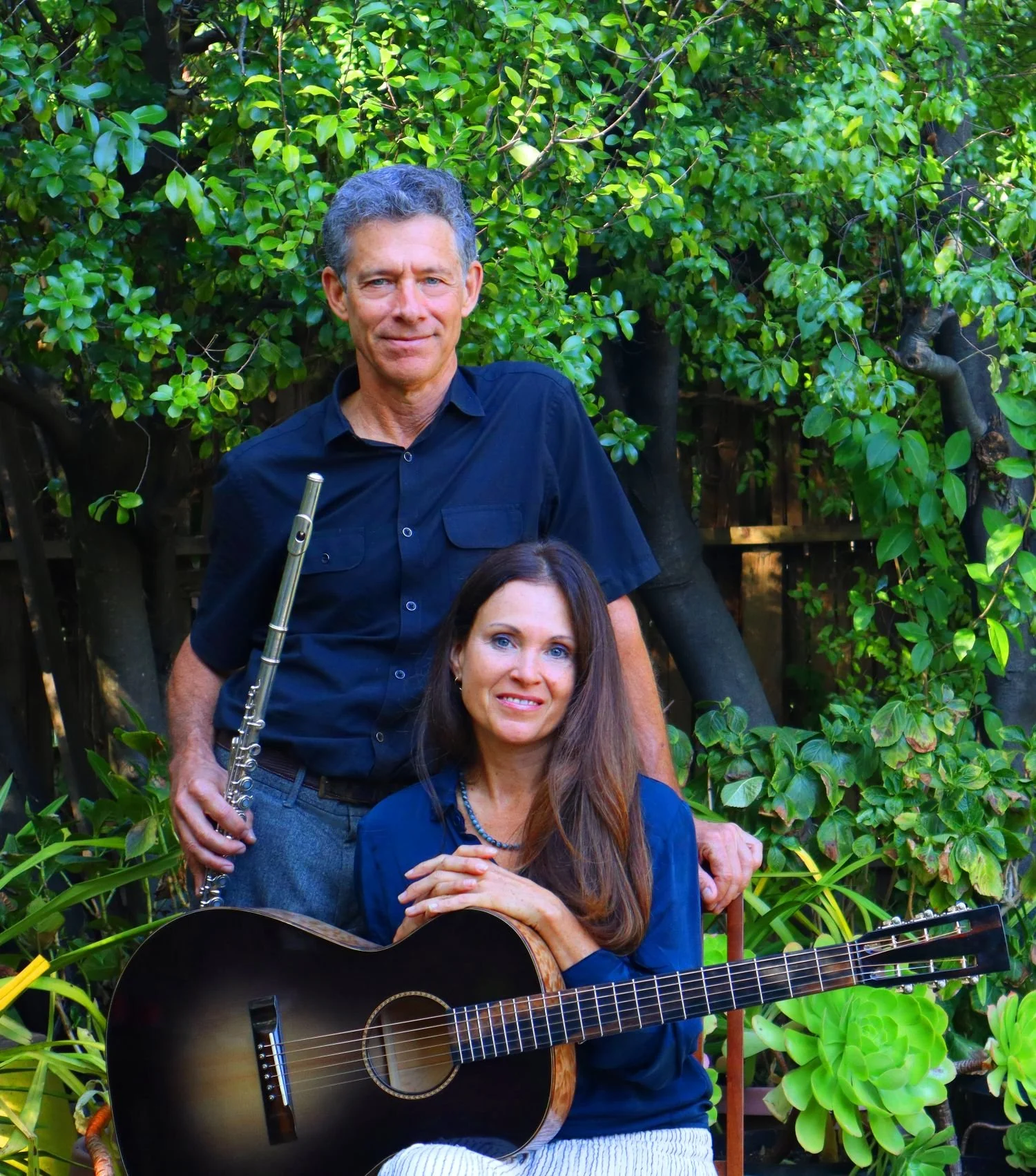 A man with gray curly hair holding a silver flute, wearing a black shirt, standing behind a seated woman with long brown hair, blue eyes, and a navy blue shirt, playing an acoustic guitar. They are outdoors in front of lush green shrubbery, with some yellow flowers visible.